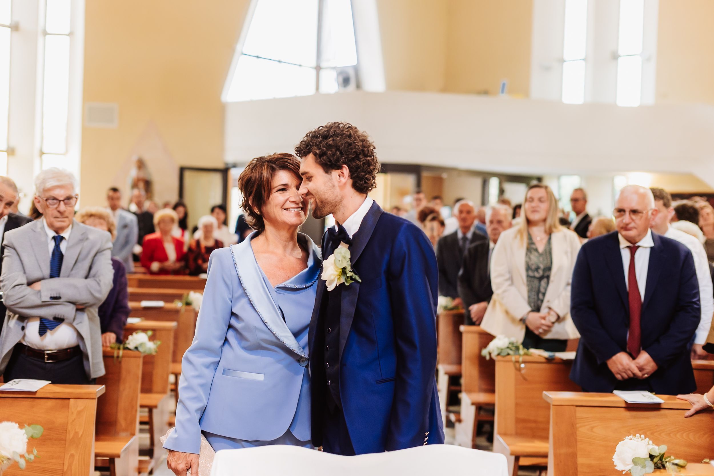 Groom in navy suit shares tender moment with woman in blue at church wedding ceremony with guests seated in pews.