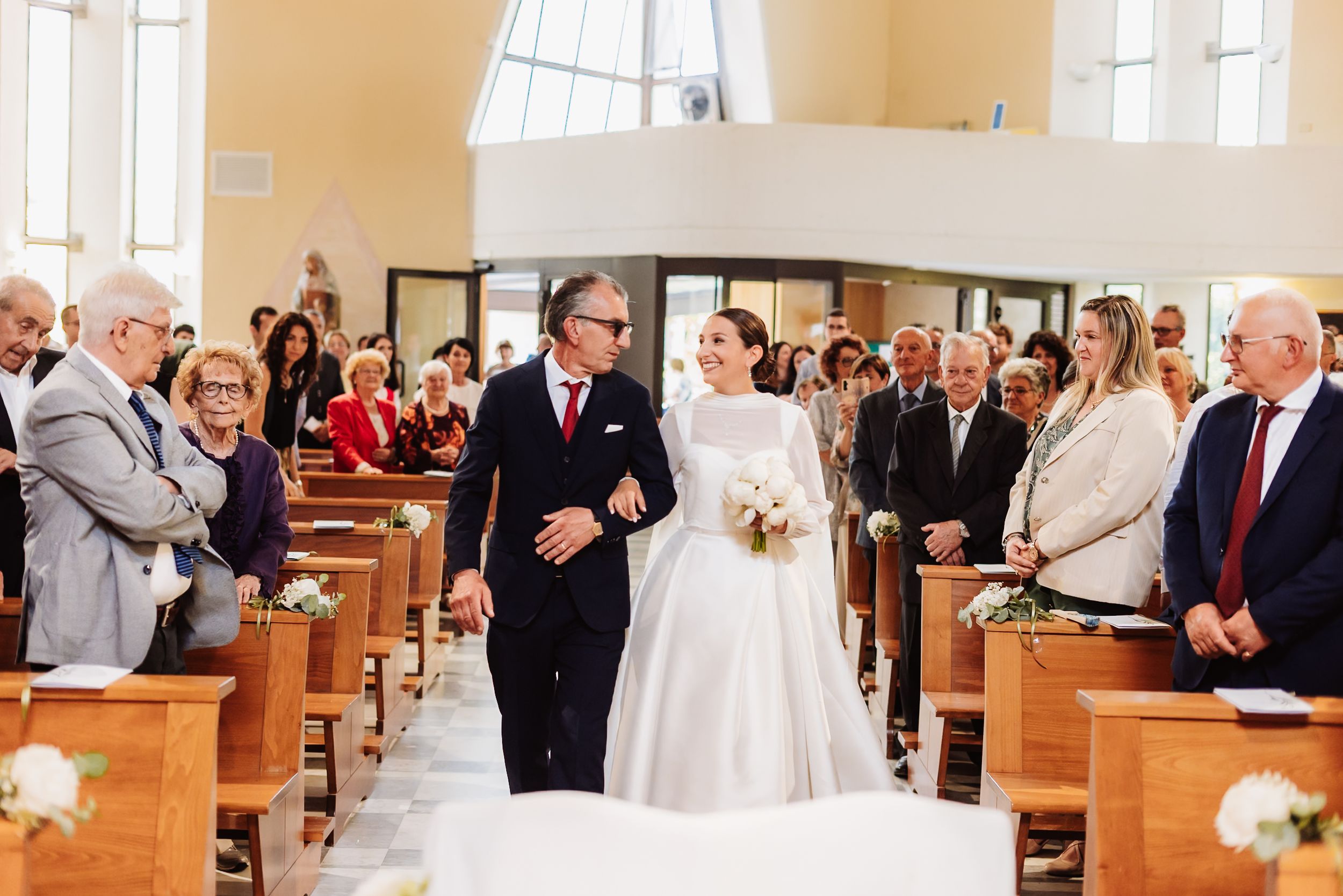 Bride walking down church aisle with escort, guests standing in wooden pews during wedding ceremony.
