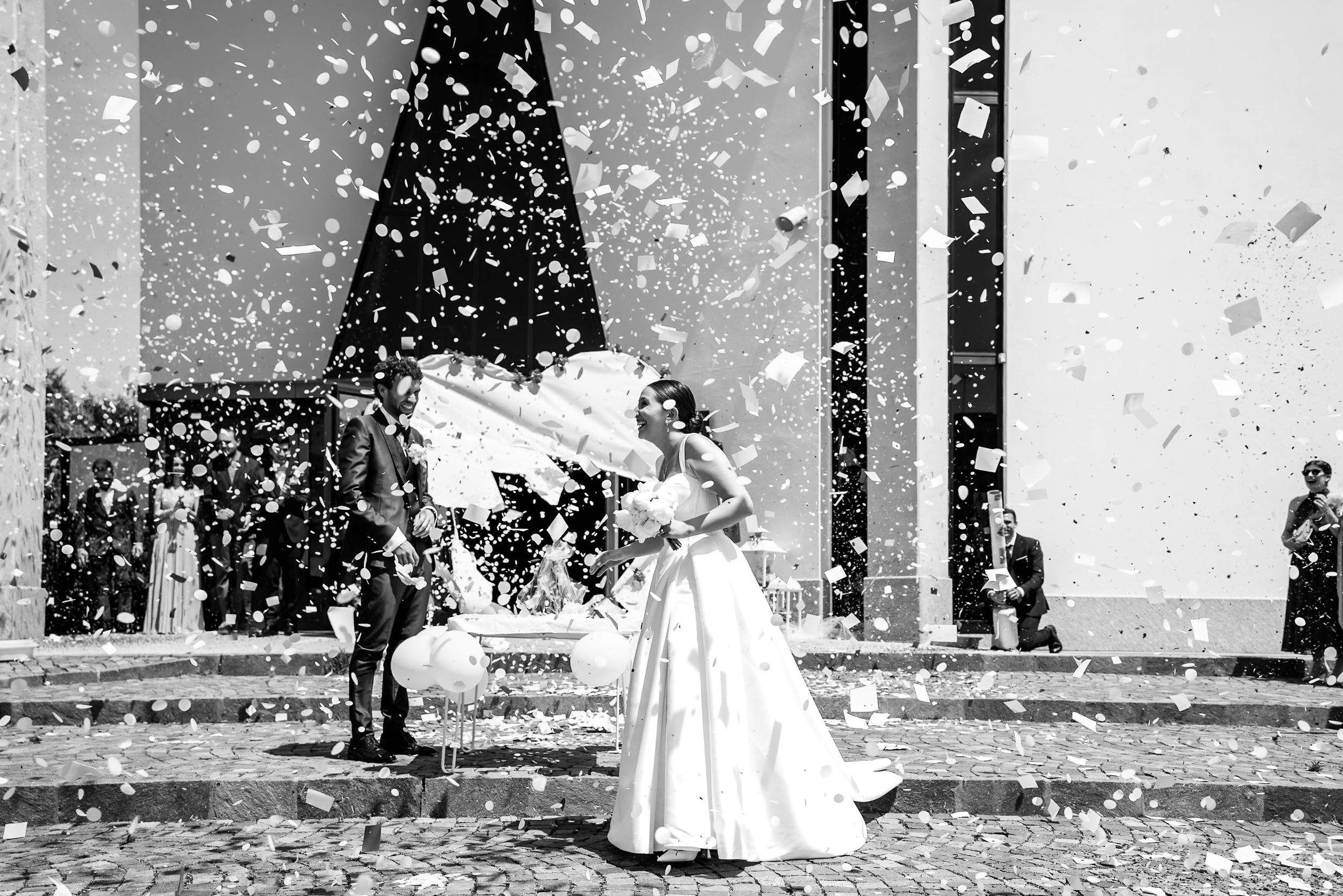 Black and white wedding photo of bride and groom celebrating with confetti outside a church.