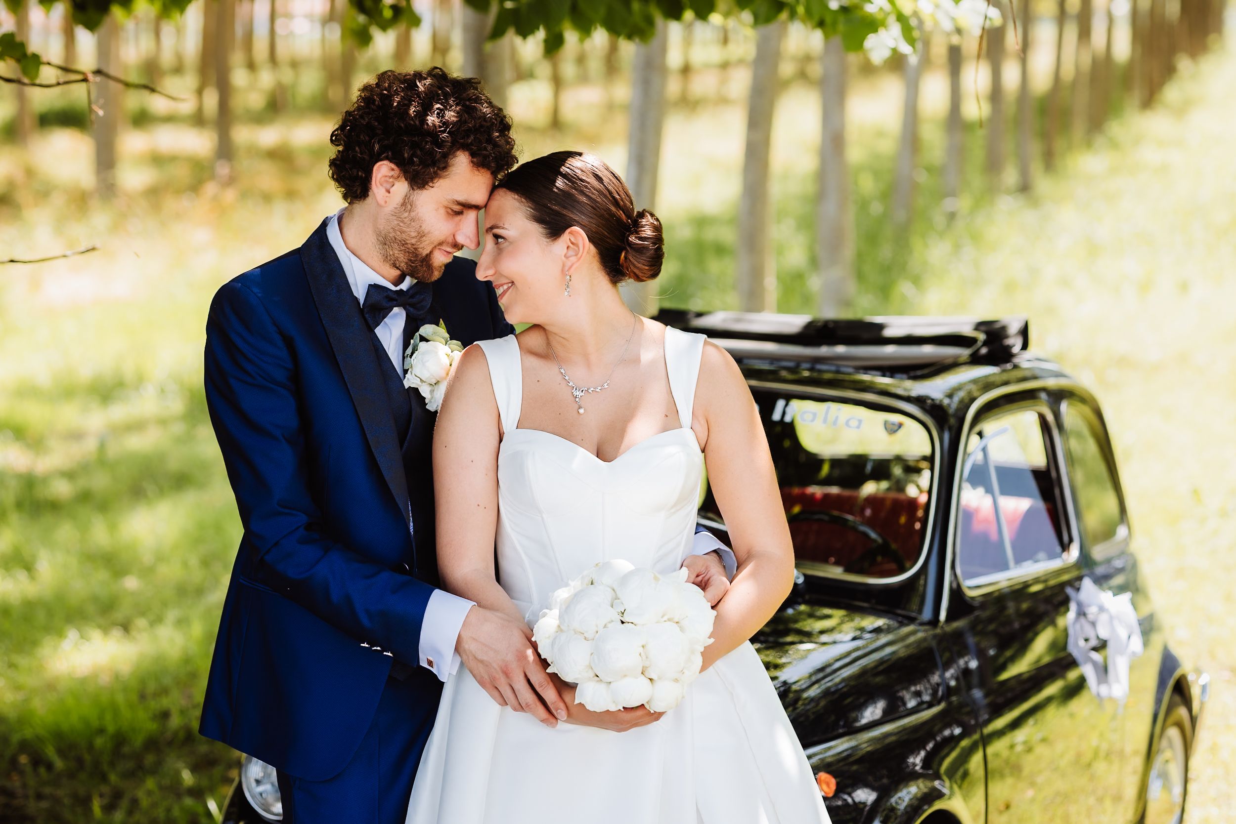 Bride and groom embracing near a vintage black car in a vineyard, bride holding white peony bouquet.