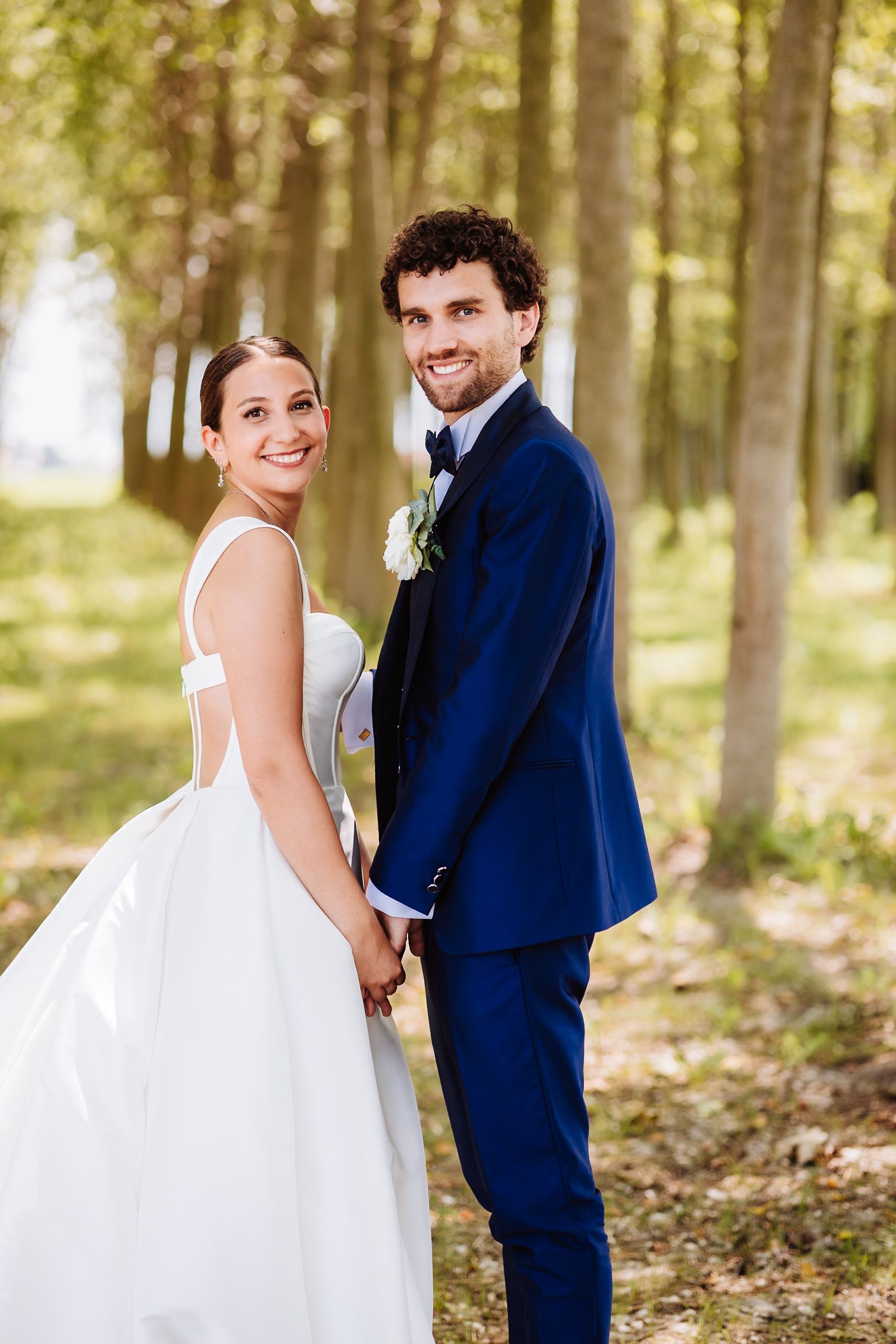 A smiling bride in a white gown and groom in a navy suit hold hands in a sunlit forest setting.