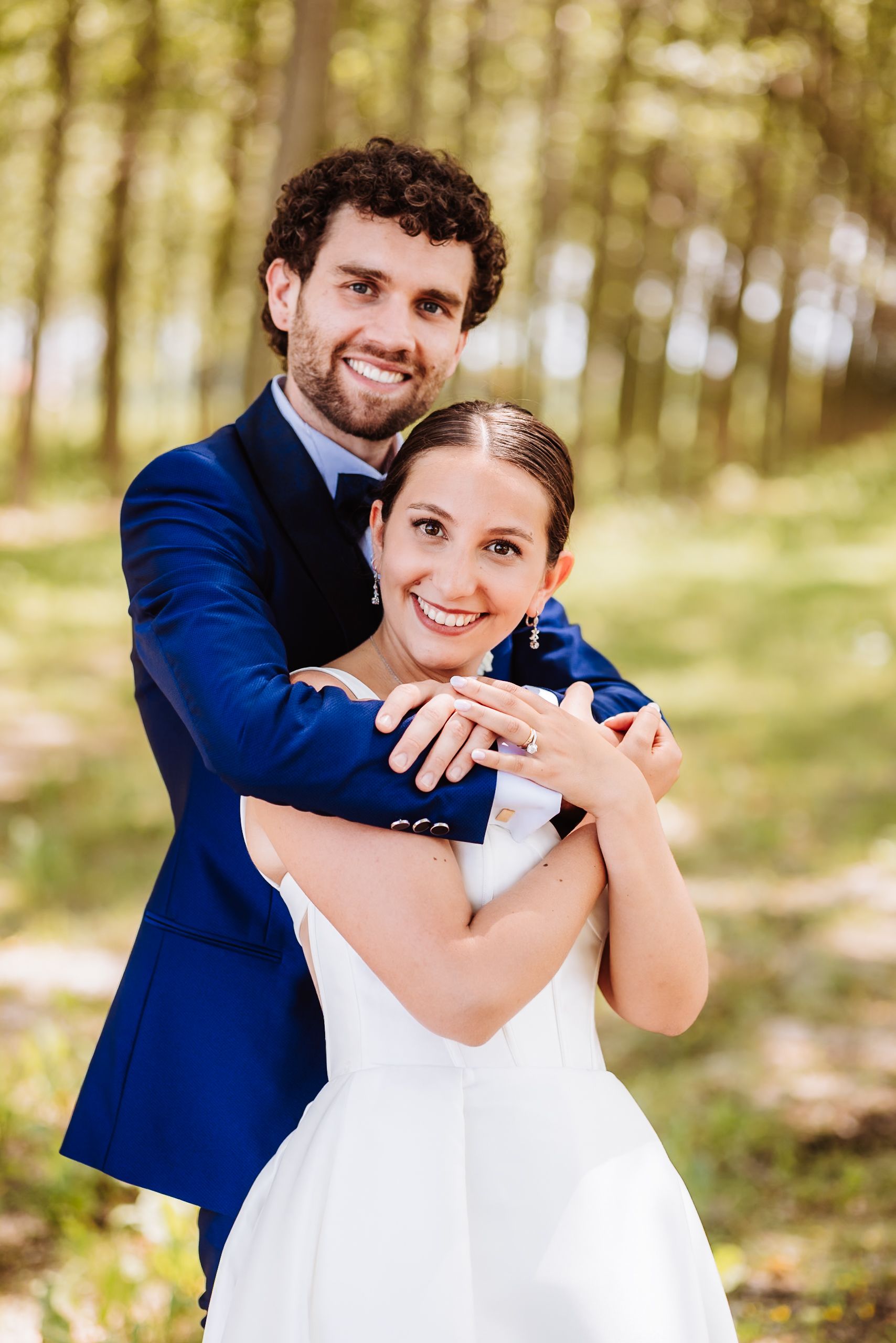 Smiling newlywed couple embracing outdoors, groom in blue suit hugging bride in white dress among trees.