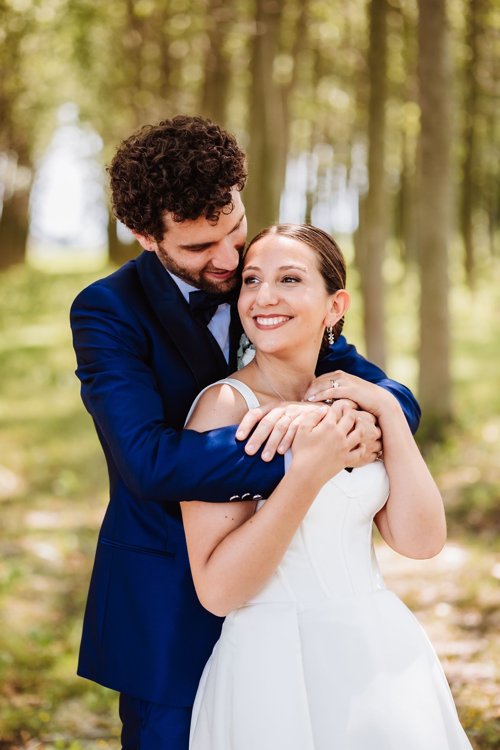 Bride and groom embrace in a sunlit forest path, she in white dress smiling brightly, he in navy suit.