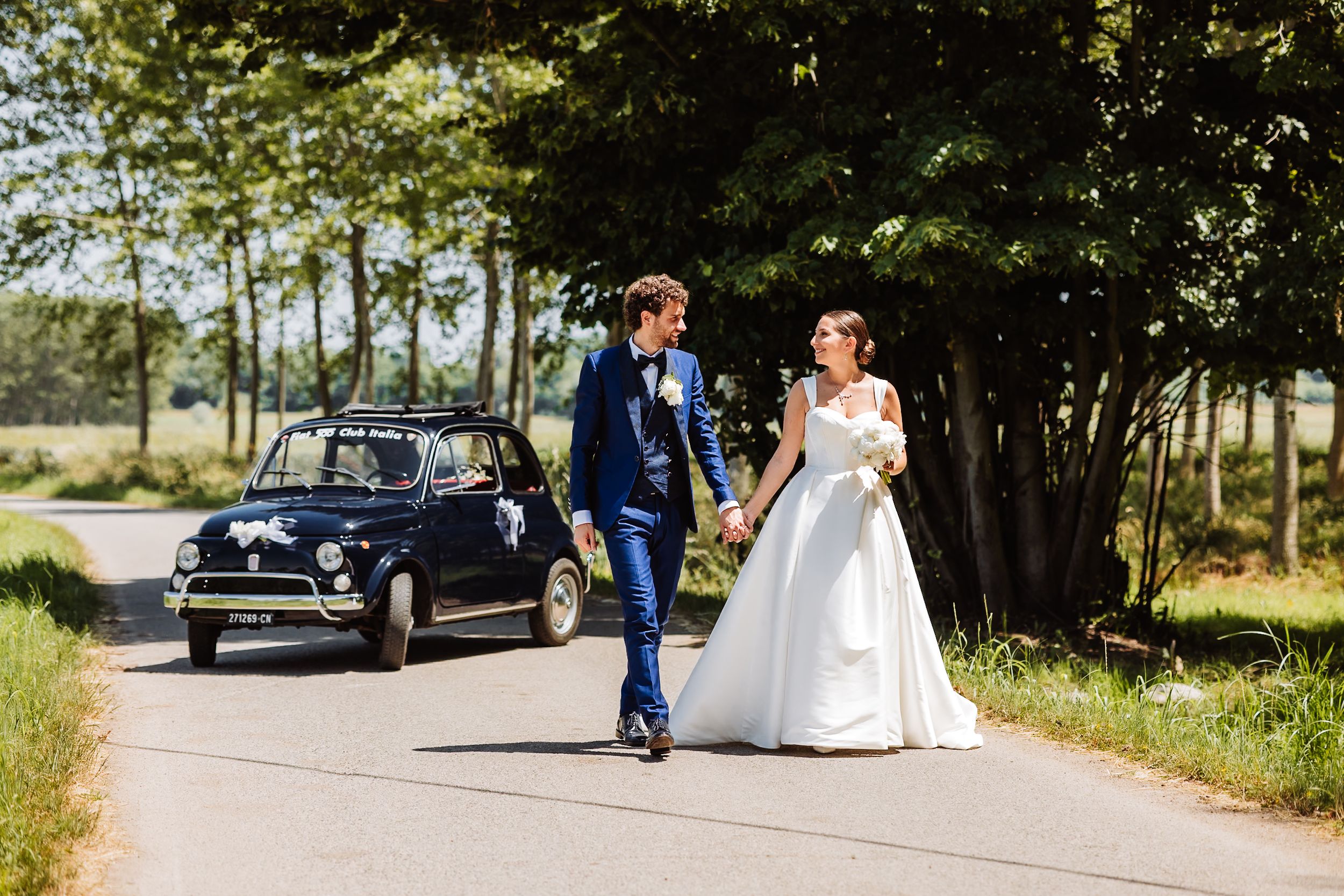 Bride and groom walking hand in hand on a country road beside a vintage navy Fiat 500 wedding car.