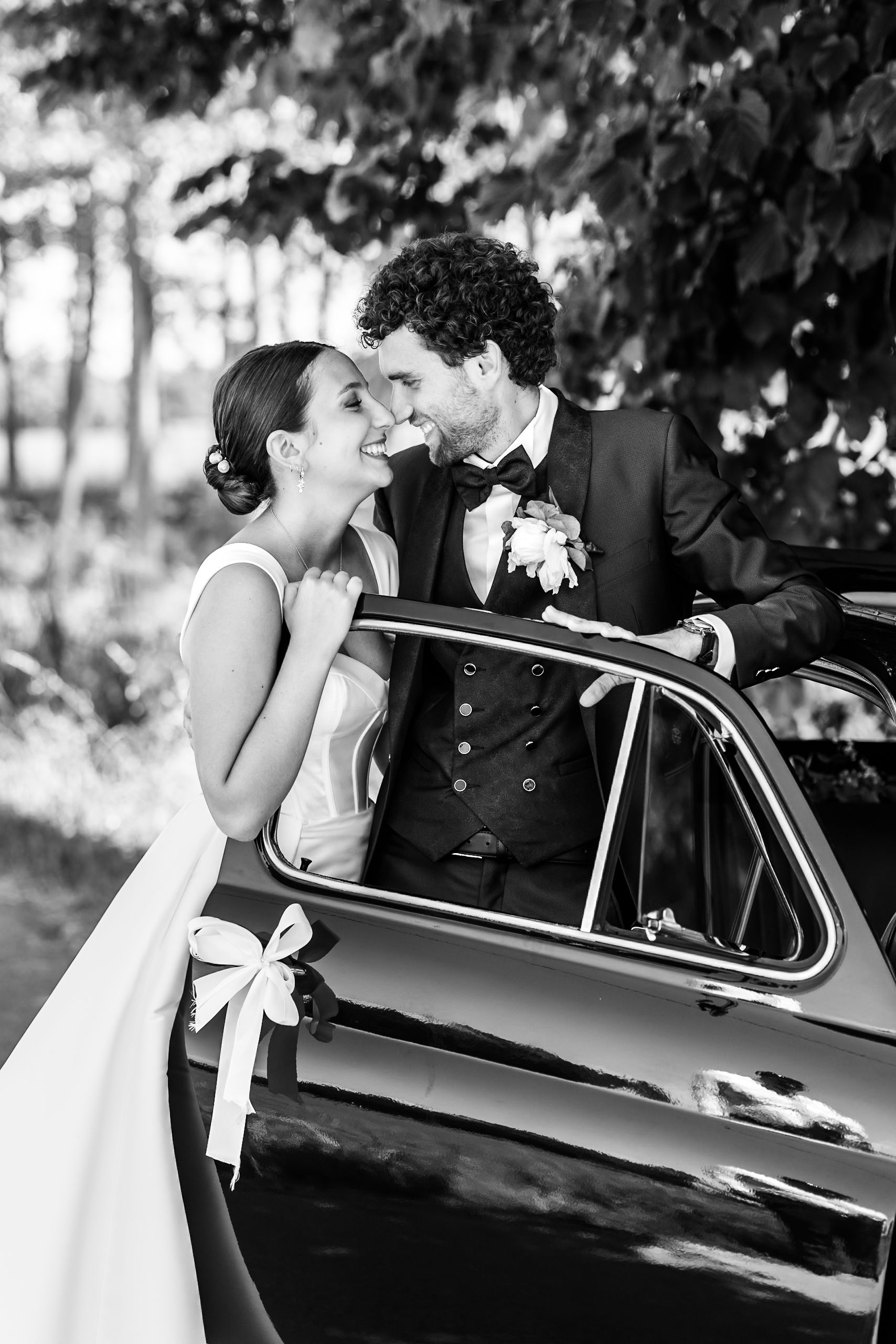 Black and white photo of a bride and groom sharing a romantic moment beside a vintage car on their wedding day.