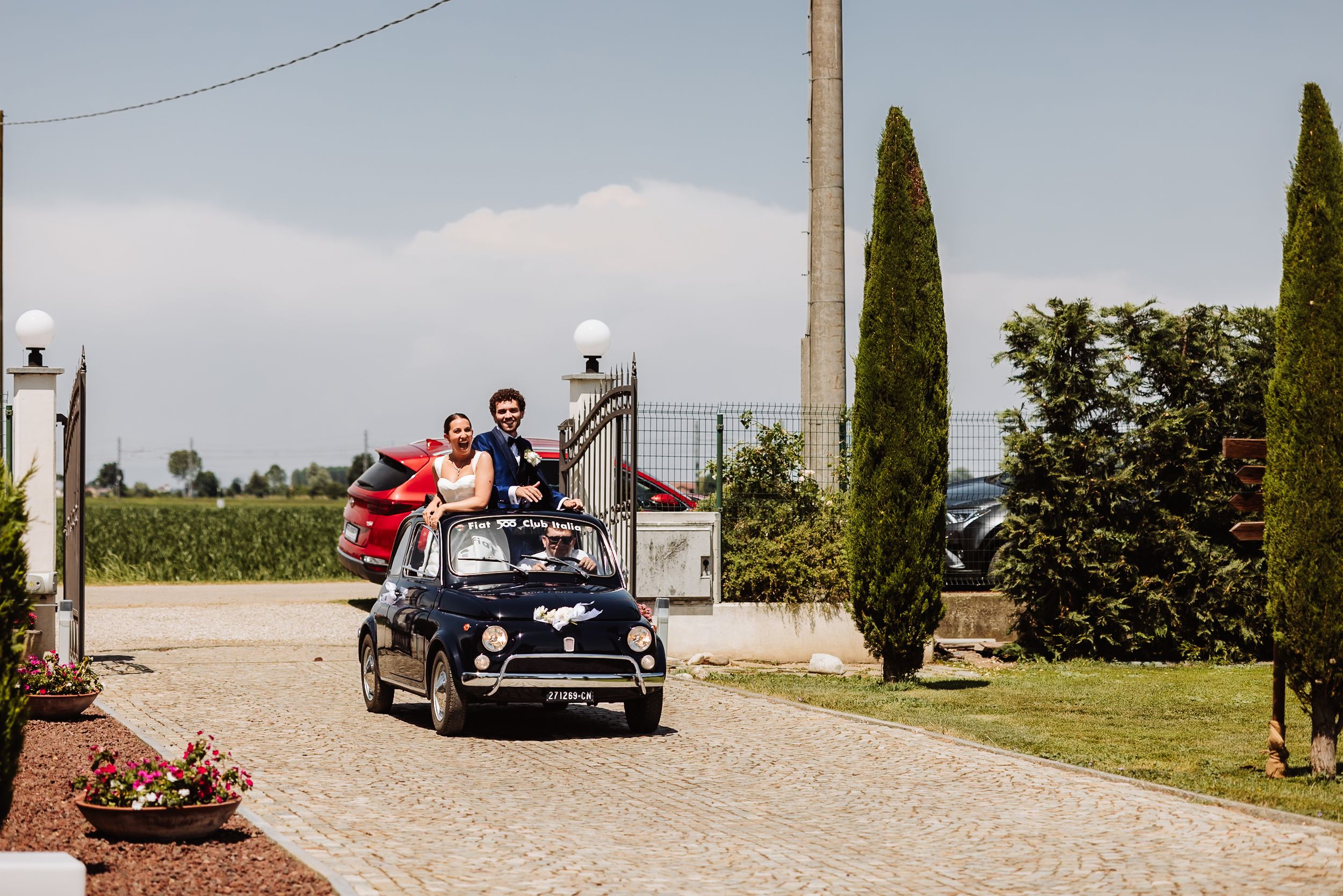 Couple standing through sunroof of vintage navy Fiat 500 driving down cobblestone road at countryside wedding venue.