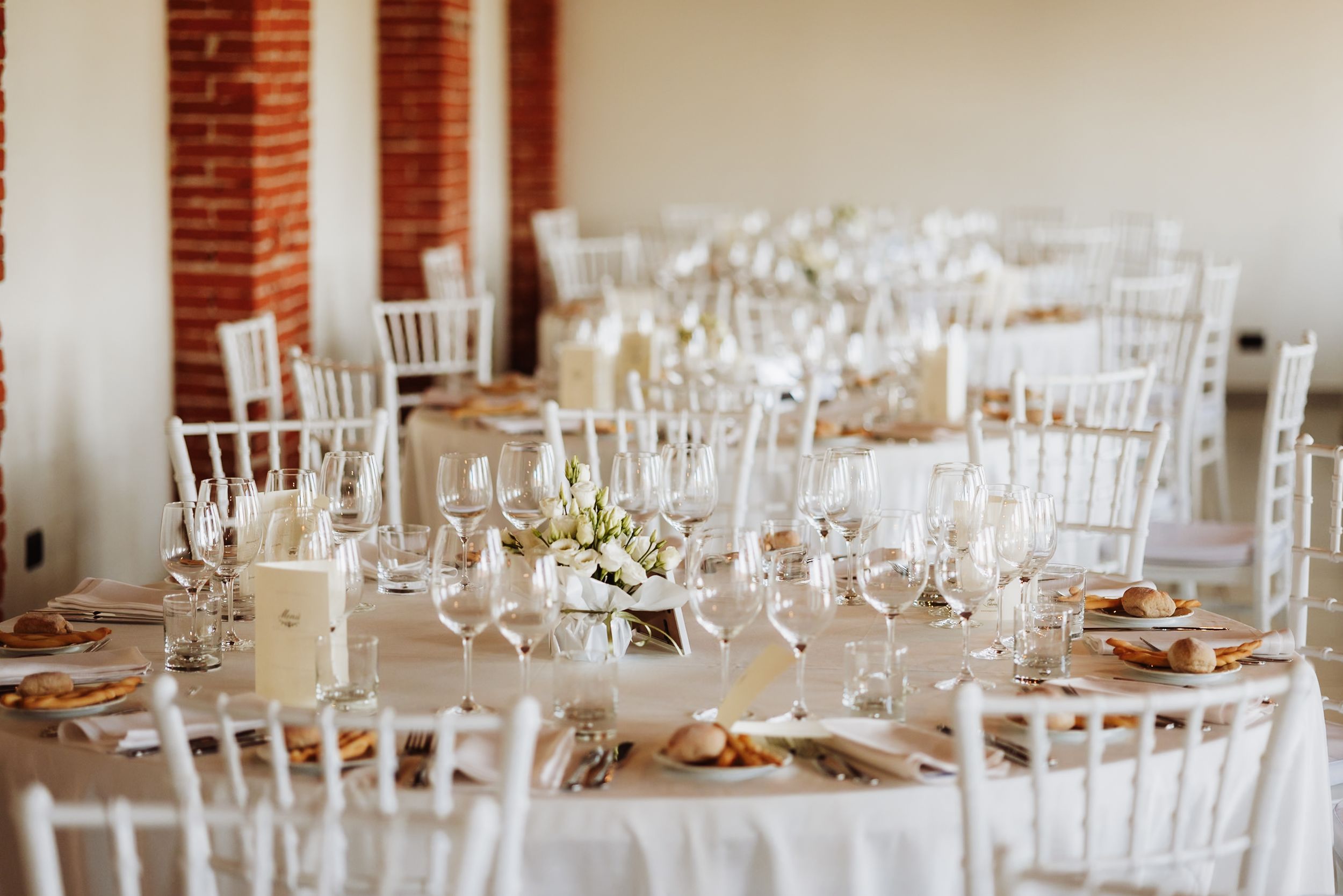 Elegant wedding reception hall with white Chiavari chairs, long tables, candles, and floral centerpieces in a bright venue.