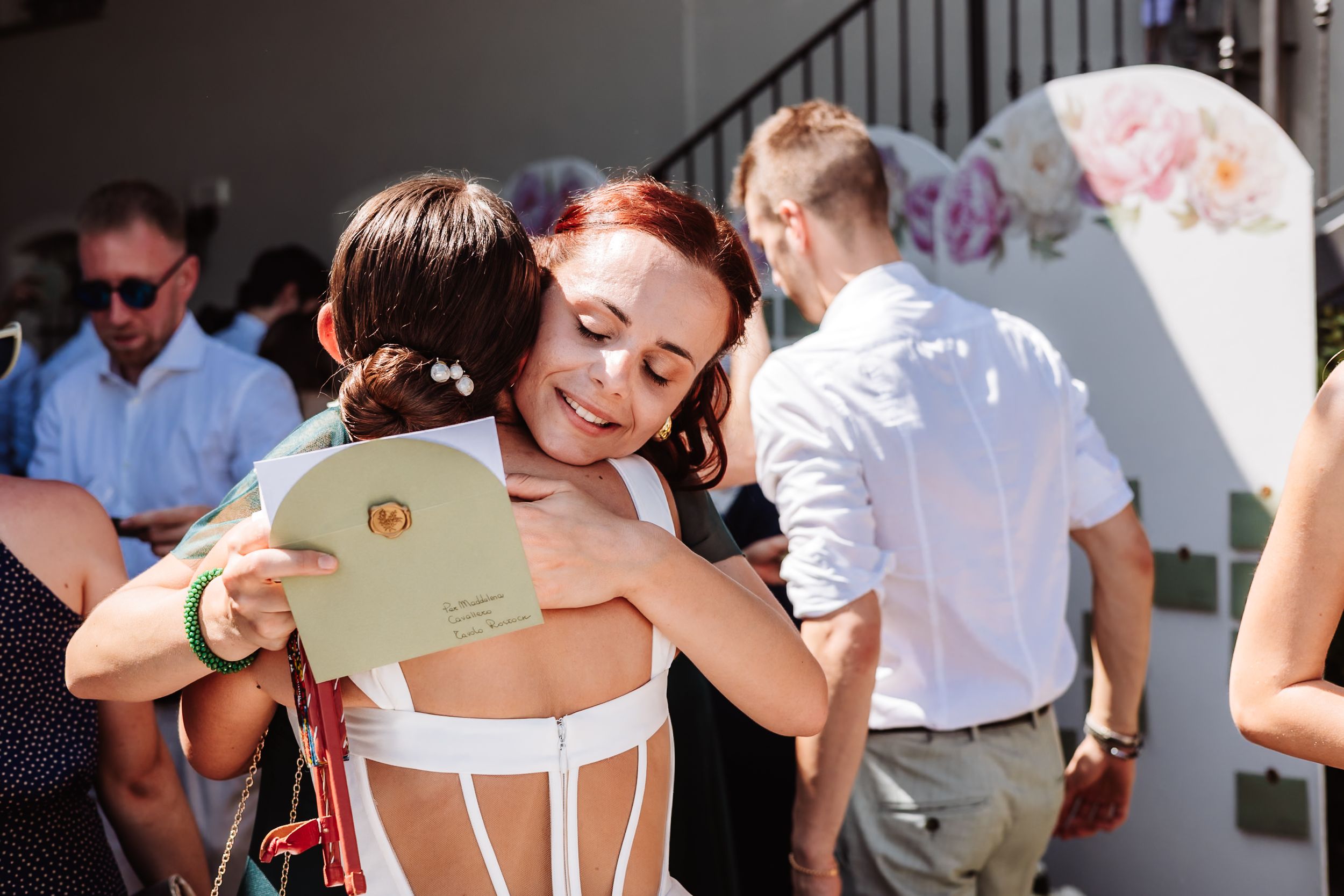 Two women share a warm hug at an outdoor wedding ceremony, one holding a floral program card.