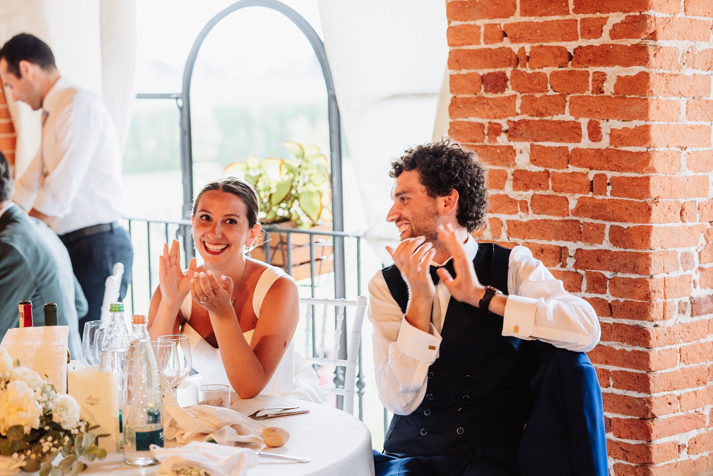 Bride and groom laughing joyfully at wedding reception table, brick wall backdrop, elegant floral centerpieces.