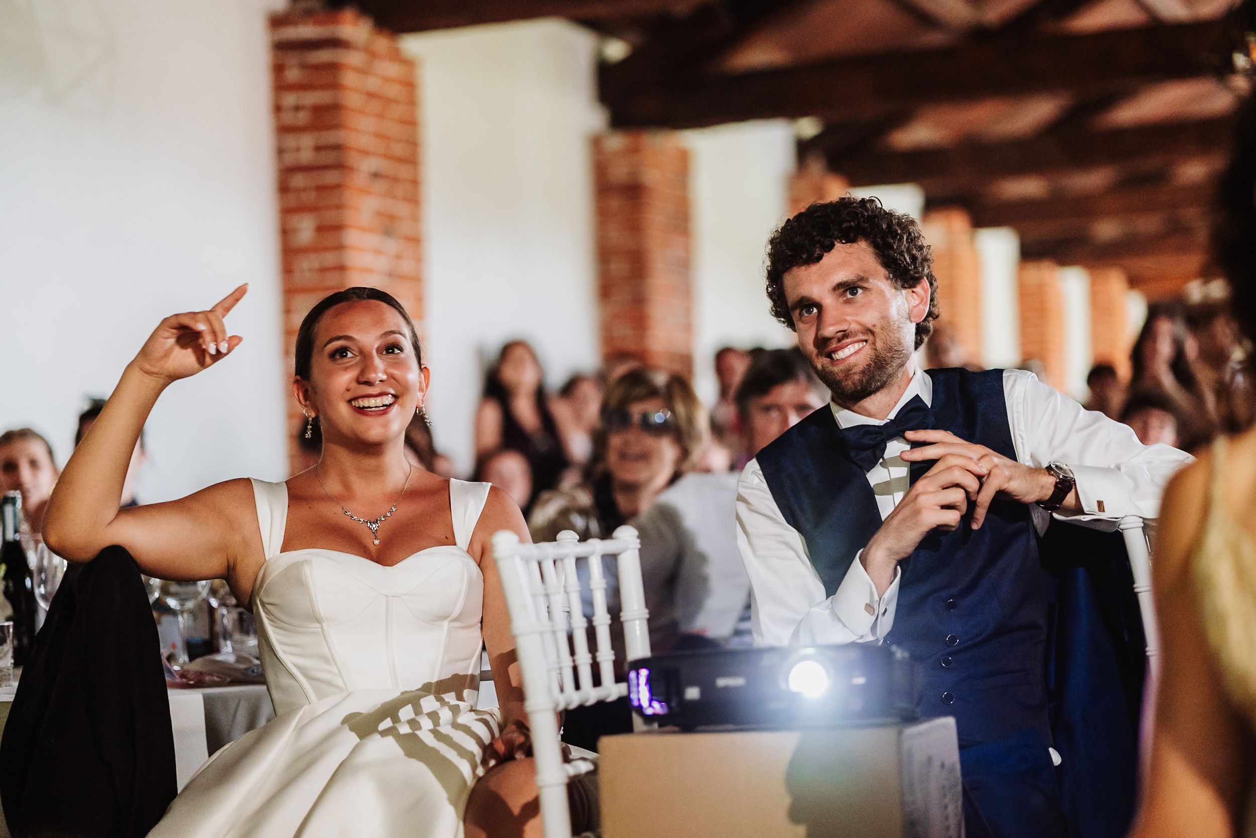 Bride in white gown and groom in navy suit laugh joyfully at wedding reception with guests in background.