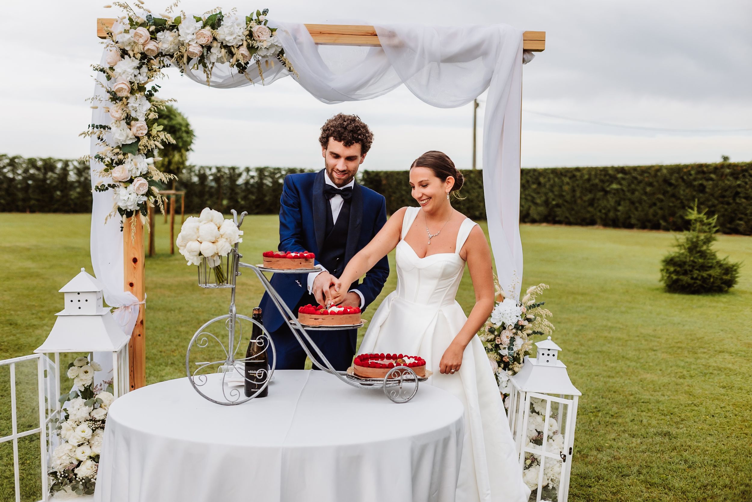 Bride and groom cutting wedding cake on decorative bicycle table under floral arch outdoors.