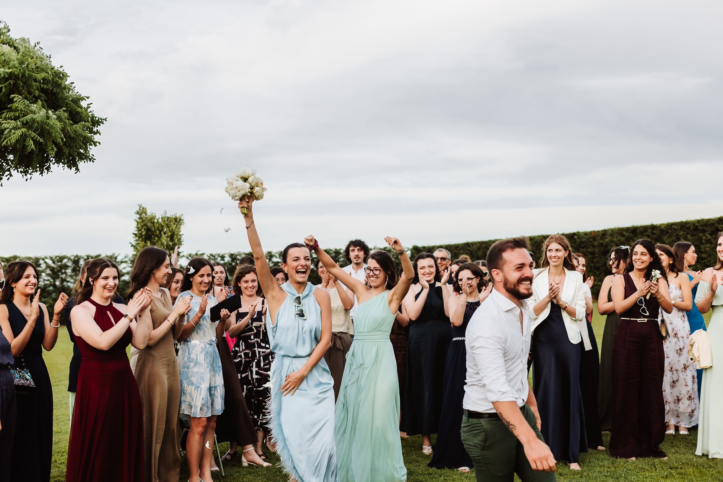 Joyful wedding guests celebrate outdoors as bride tosses bouquet, with crowd clapping and cheering on green lawn.