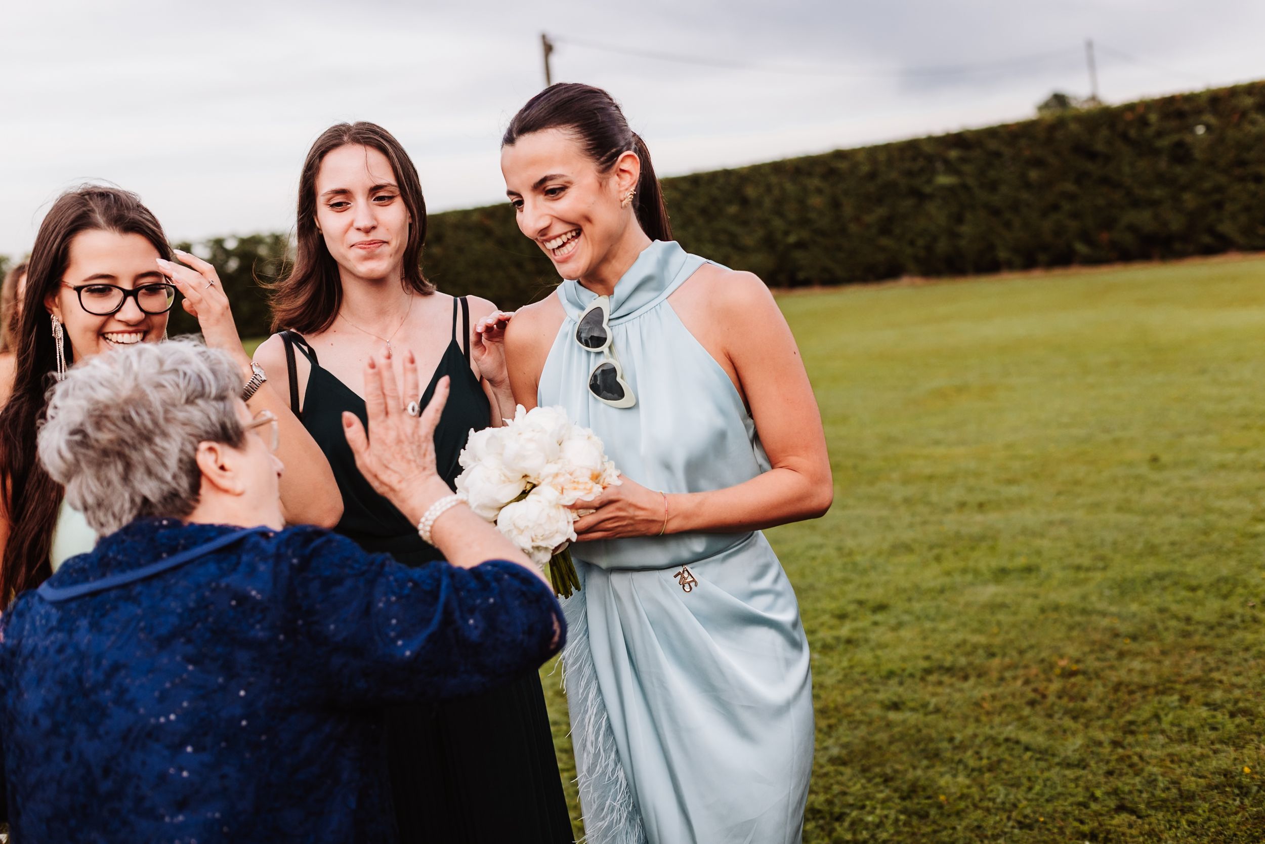 Women in elegant attire gather outdoors, one holding white flowers, sharing a joyful moment at a formal event.