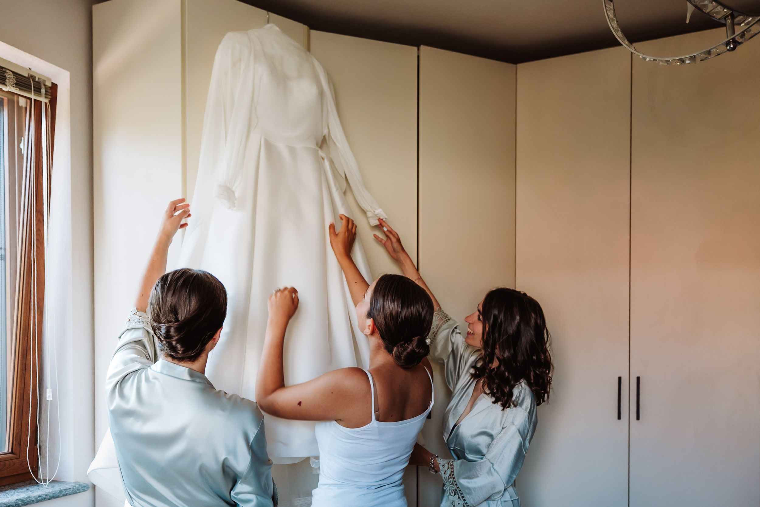 Three women in satin robes admire a white wedding dress hanging on a wardrobe in a bright, elegant room.