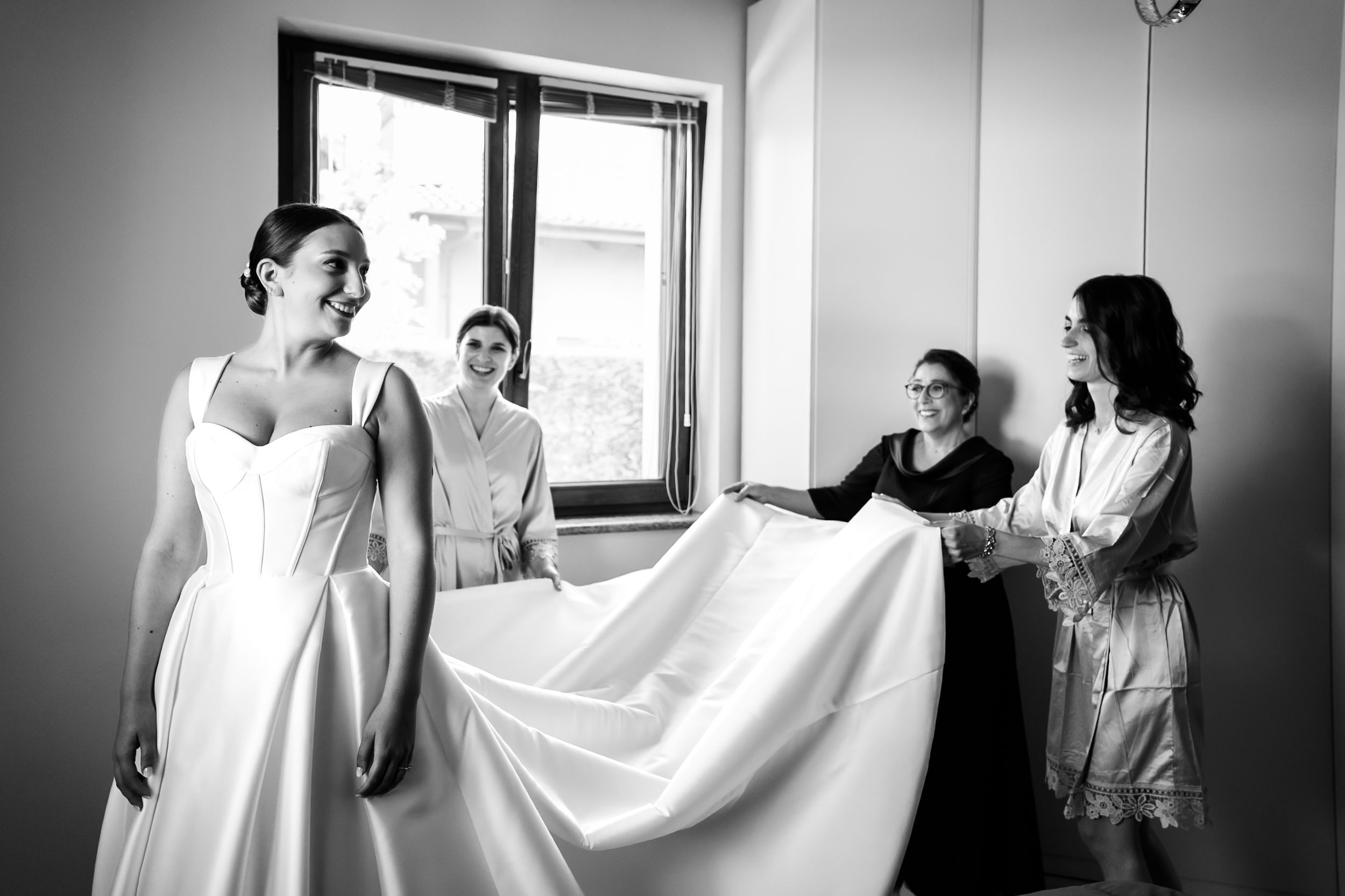 Bride in white gown assisted by bridesmaids adjusting her train in a bright room, black and white wedding photo.