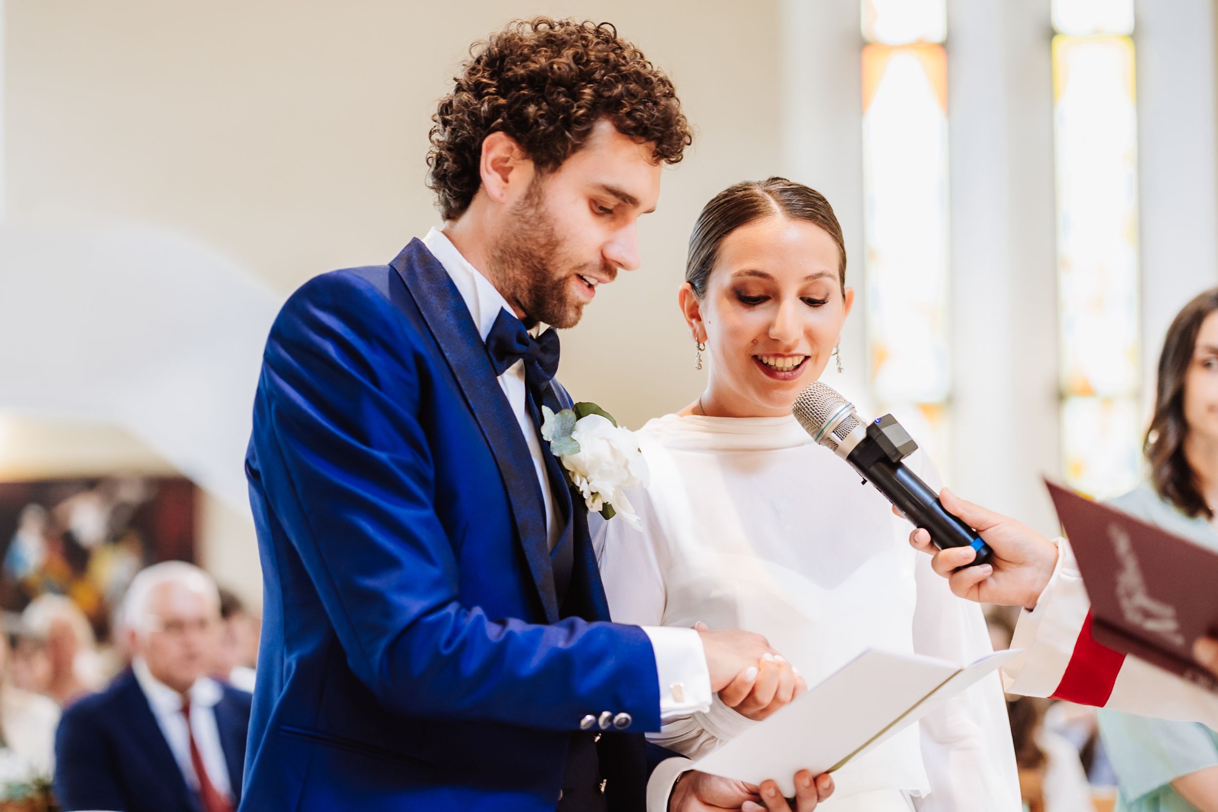 Bride and groom sharing vows at church altar, groom in blue suit holding paper near microphone.