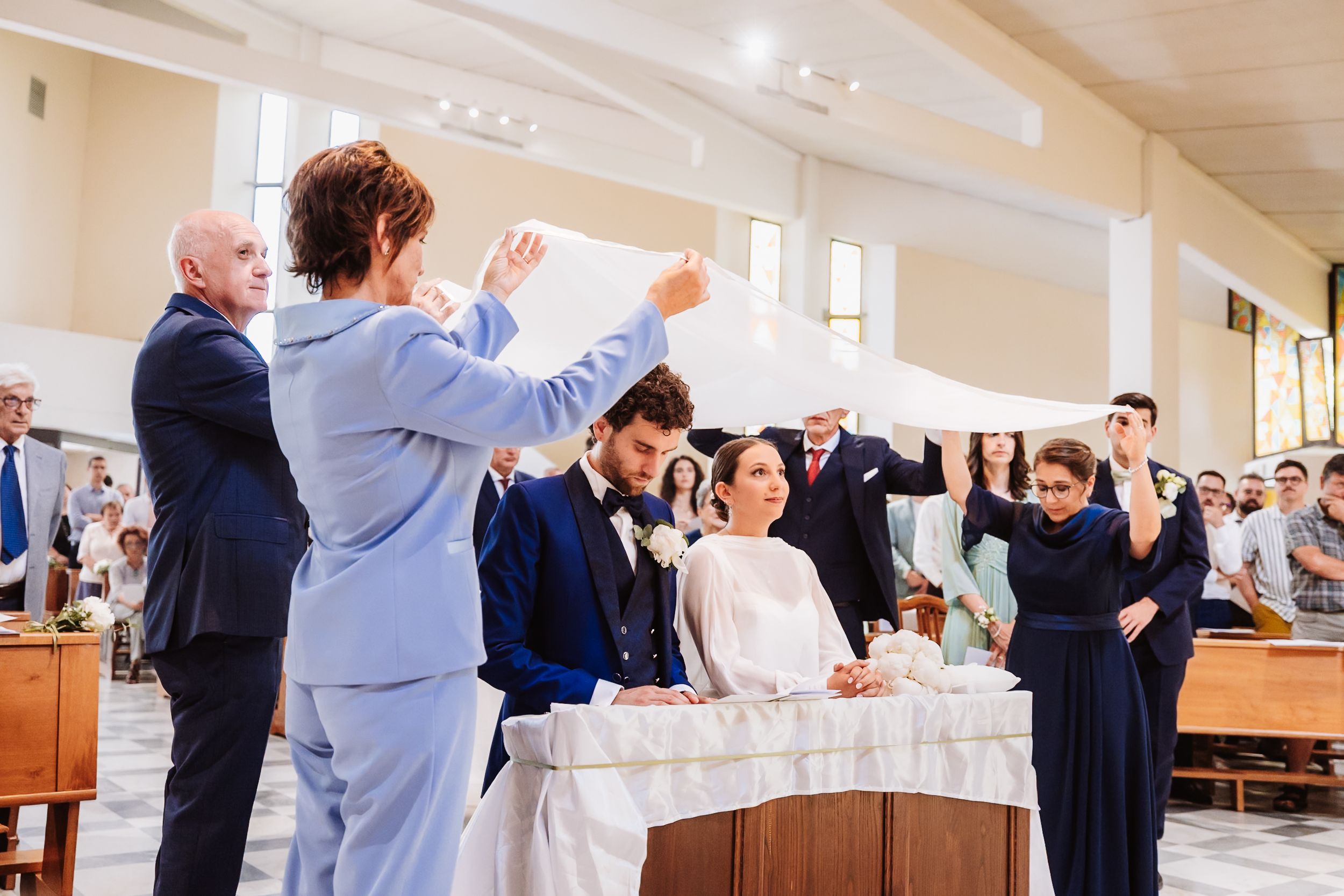 Wedding ceremony in a church with bride and groom seated as guests hold a white veil over them.