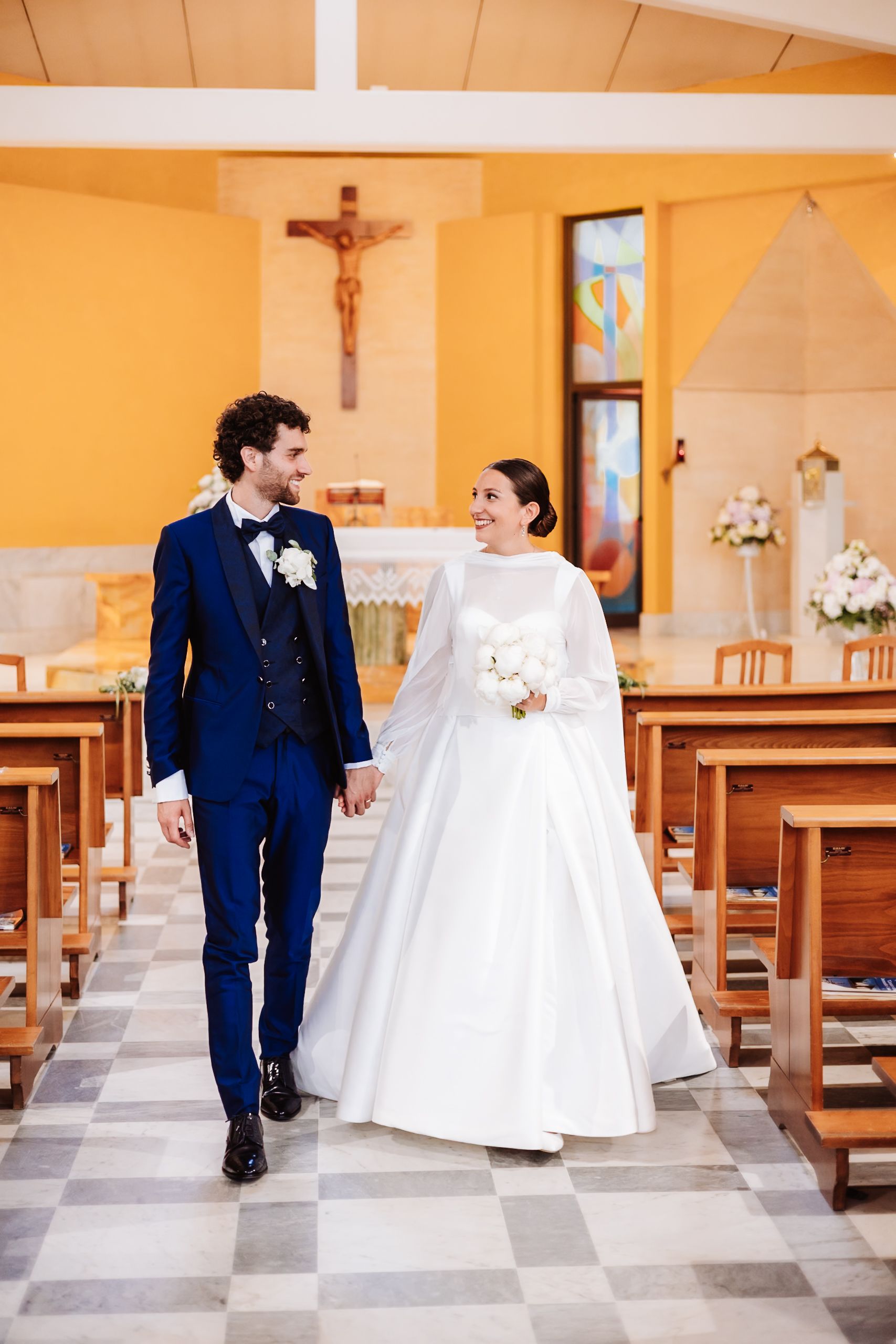 Bride and groom walk hand in hand down church aisle, she in white gown with bouquet, he in navy suit.