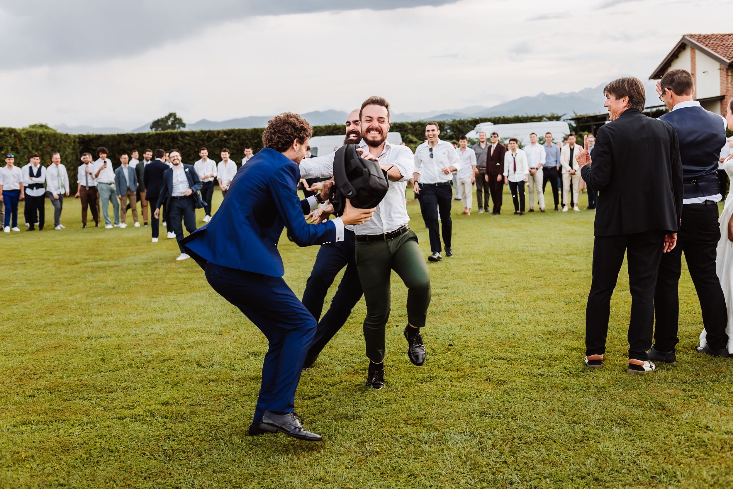 Two men playfully wrestling on green lawn at outdoor wedding reception with guests watching in background.