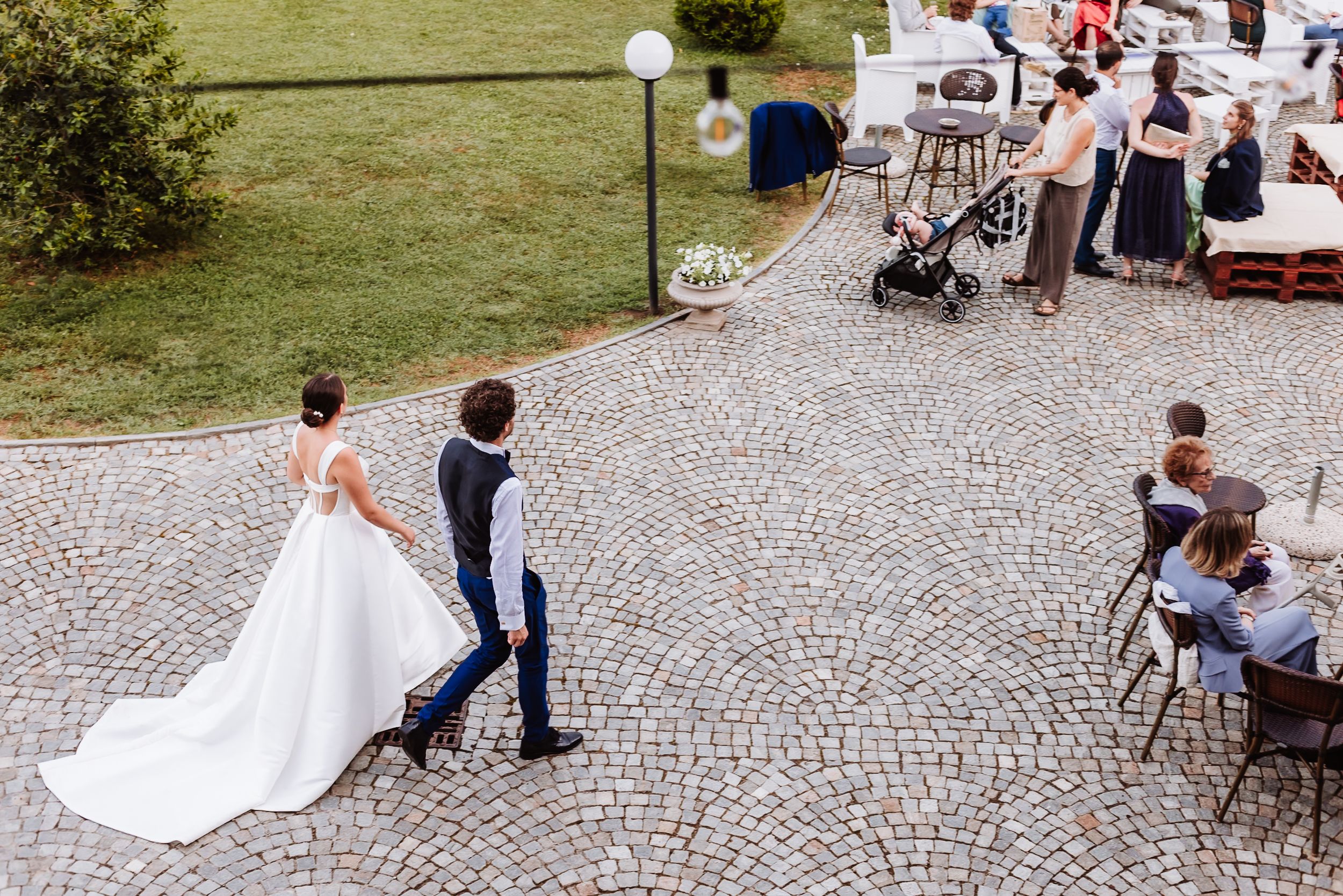 Bride and groom walking hand in hand on cobblestone plaza at outdoor wedding reception with guests seated nearby.