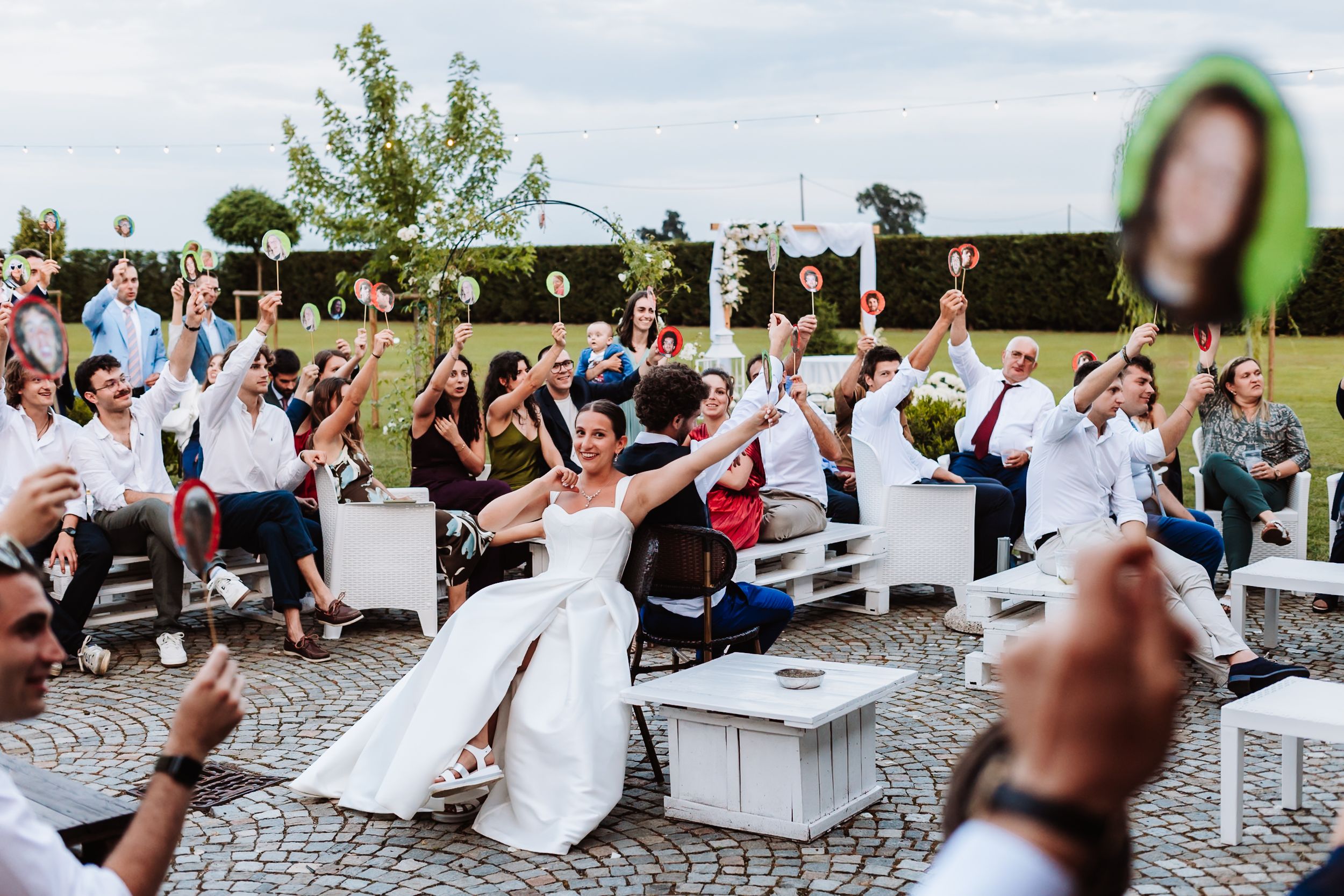 Bride in white dress celebrates with wedding guests raising hands outdoors at a garden reception ceremony.