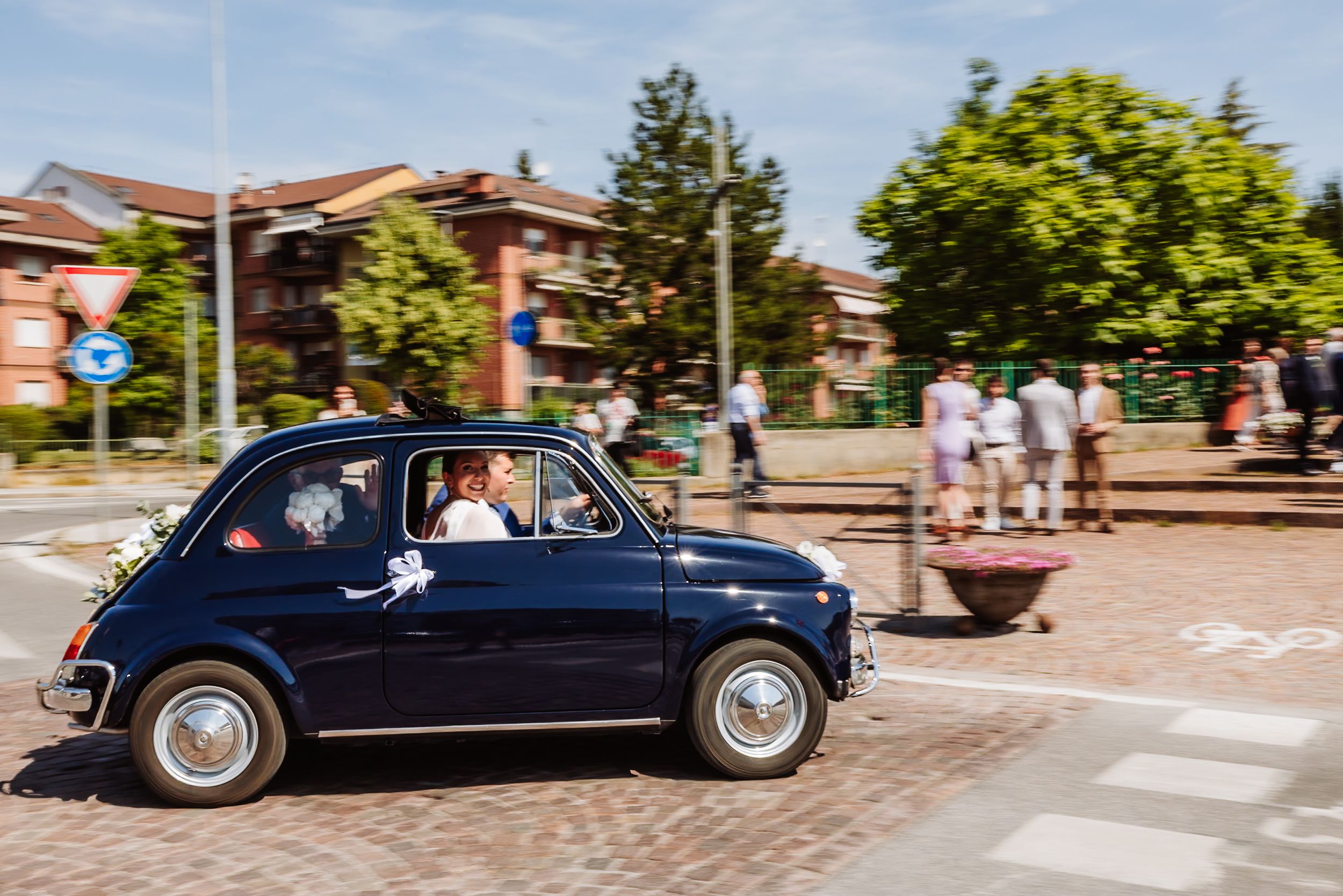 Vintage blue Fiat 500 carrying newlyweds drives through a sunny Italian street on a wedding day.