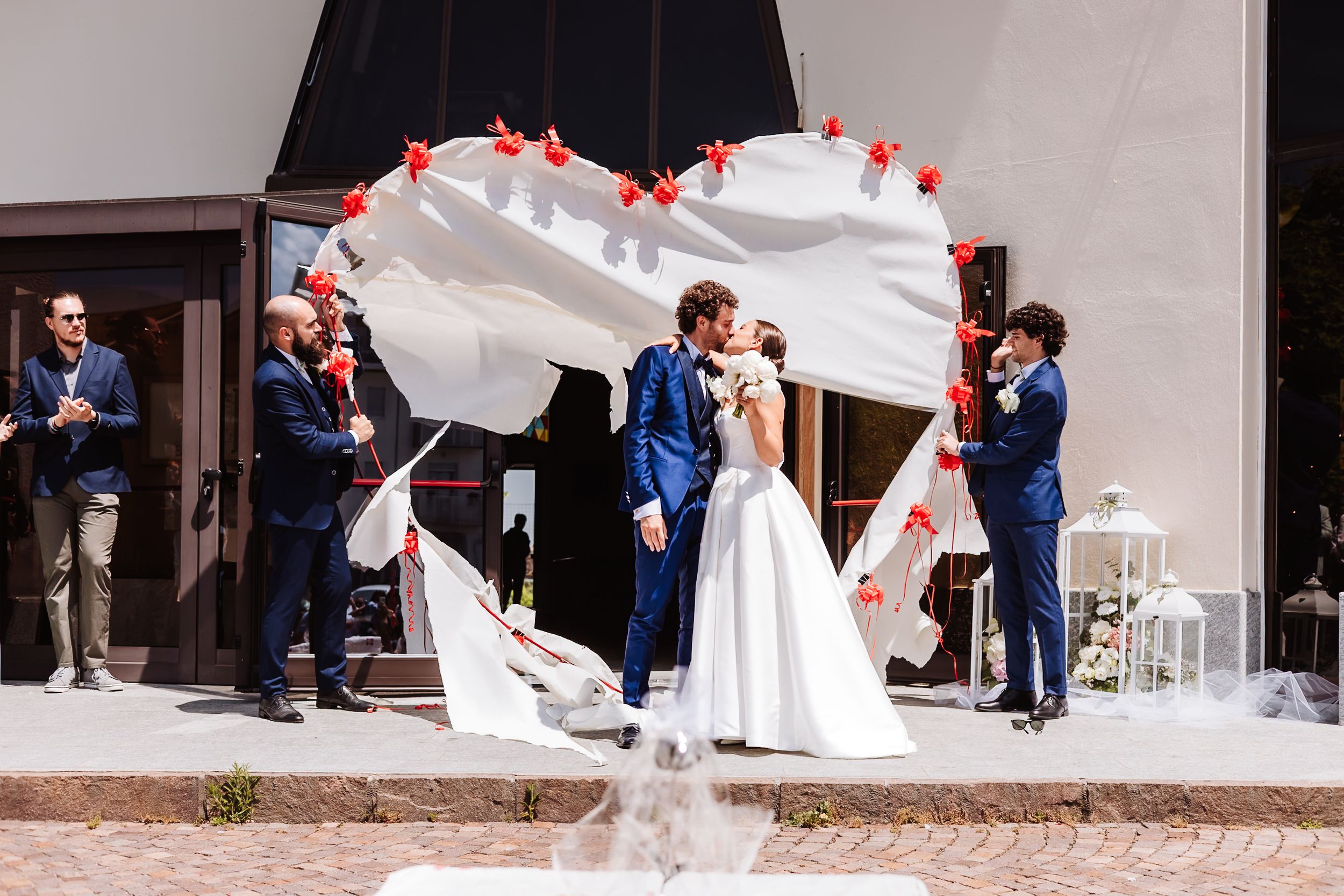Bride and groom share a kiss under a white floral arch with red accents at an outdoor wedding ceremony.