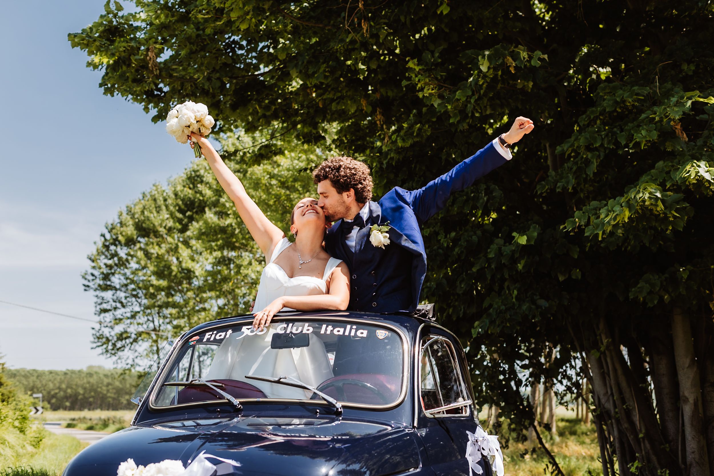 Joyful newlyweds celebrate atop a vintage navy blue wedding car, bride holding bouquet aloft on a sunny day.
