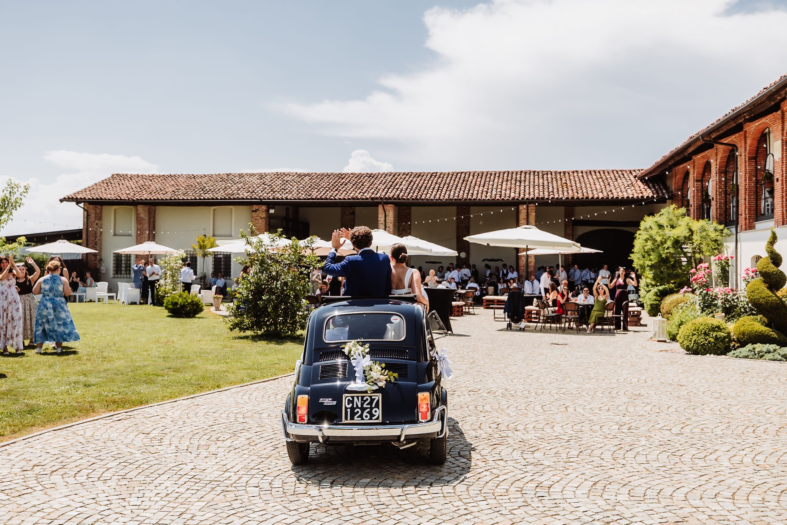 Newlyweds ride in a vintage Fiat 500 through cobblestone courtyard at elegant Italian villa wedding venue.