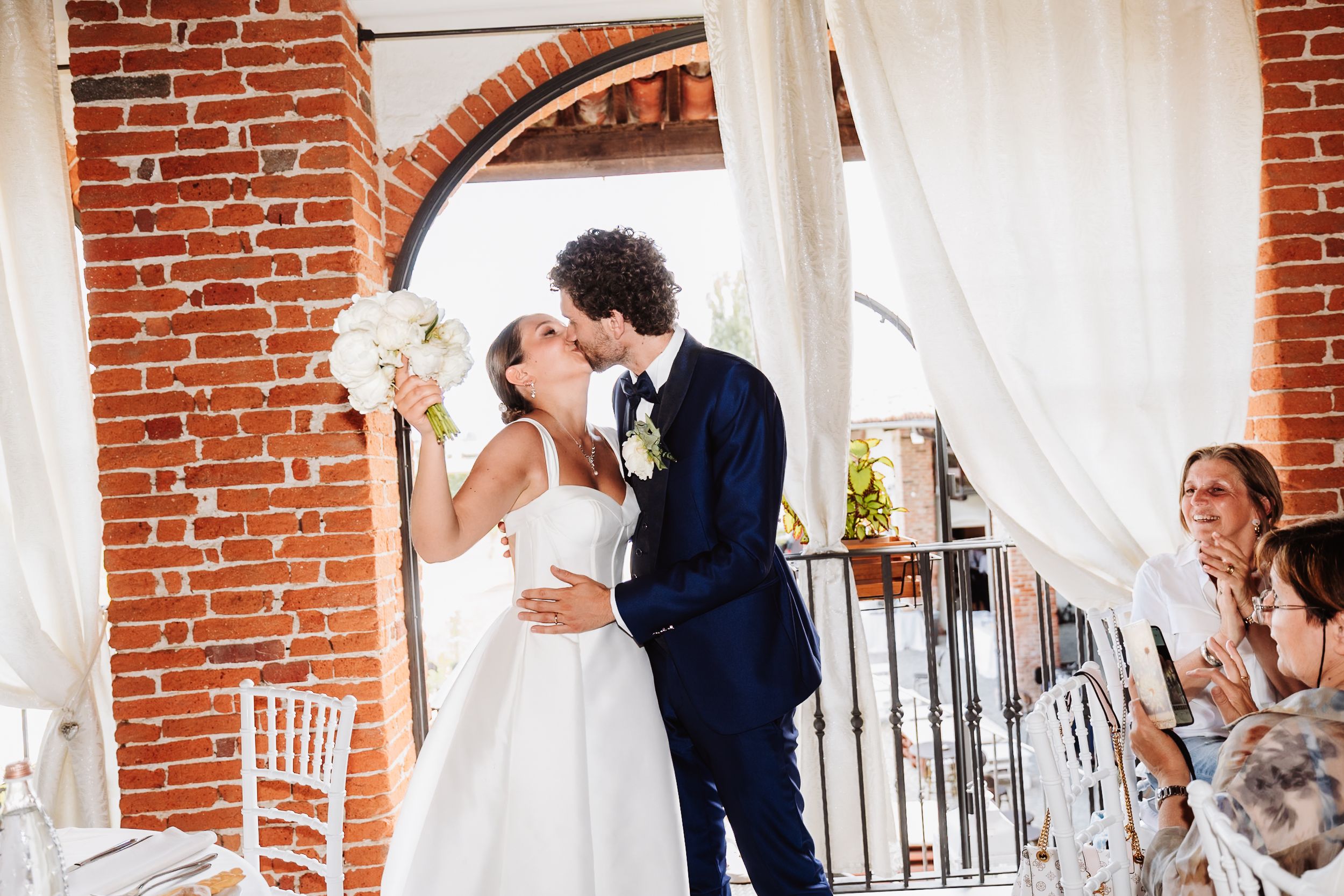 Bride and groom share a romantic kiss at their wedding reception in an elegant brick-arched venue with white drapes.
