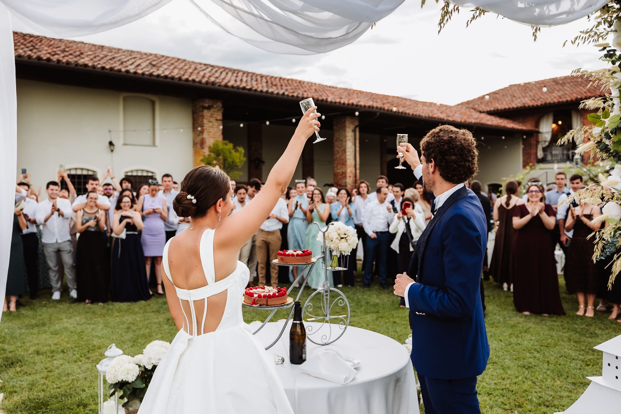 Bride and groom celebrating with raised glasses at outdoor wedding reception surrounded by applauding guests.