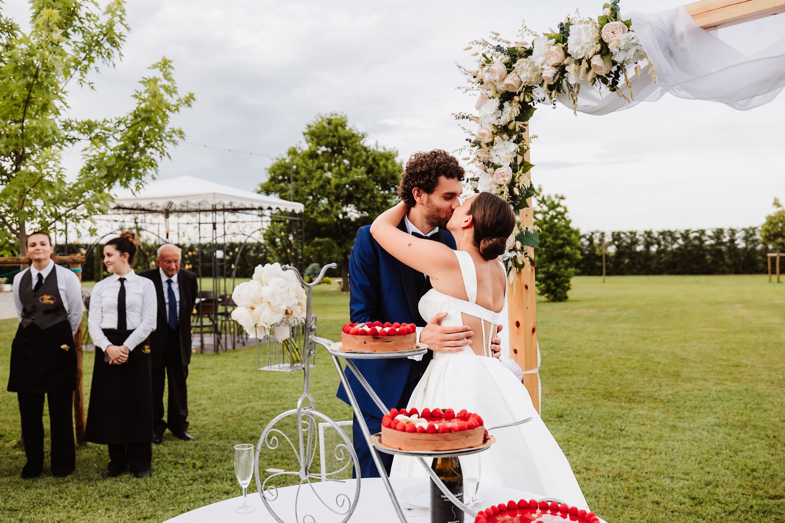 Bride and groom share a kiss outdoors at wedding ceremony with floral arch and decorated cake display on green lawn.
