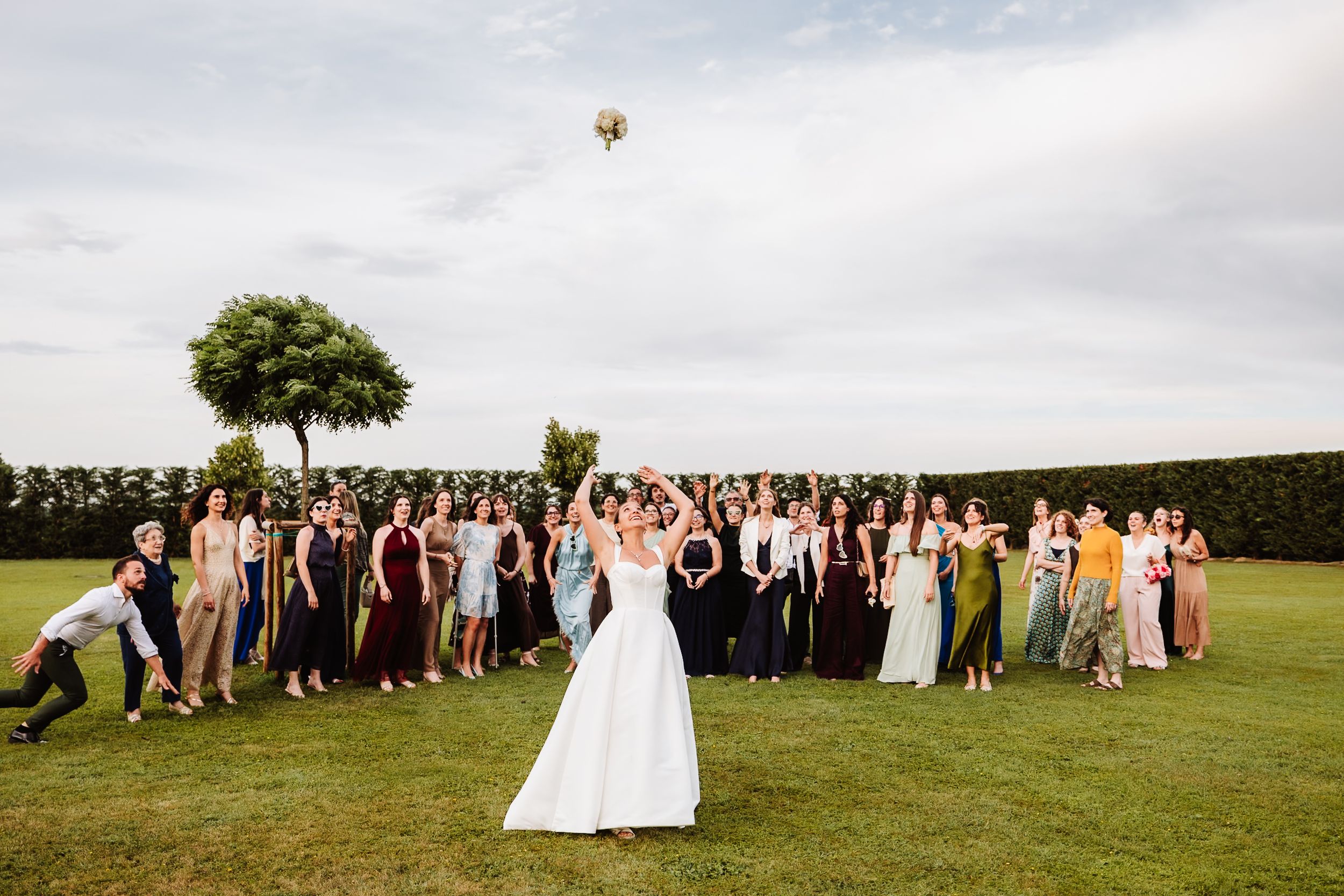 Bride tosses bouquet to guests lined up on green lawn at outdoor wedding reception under cloudy sky.