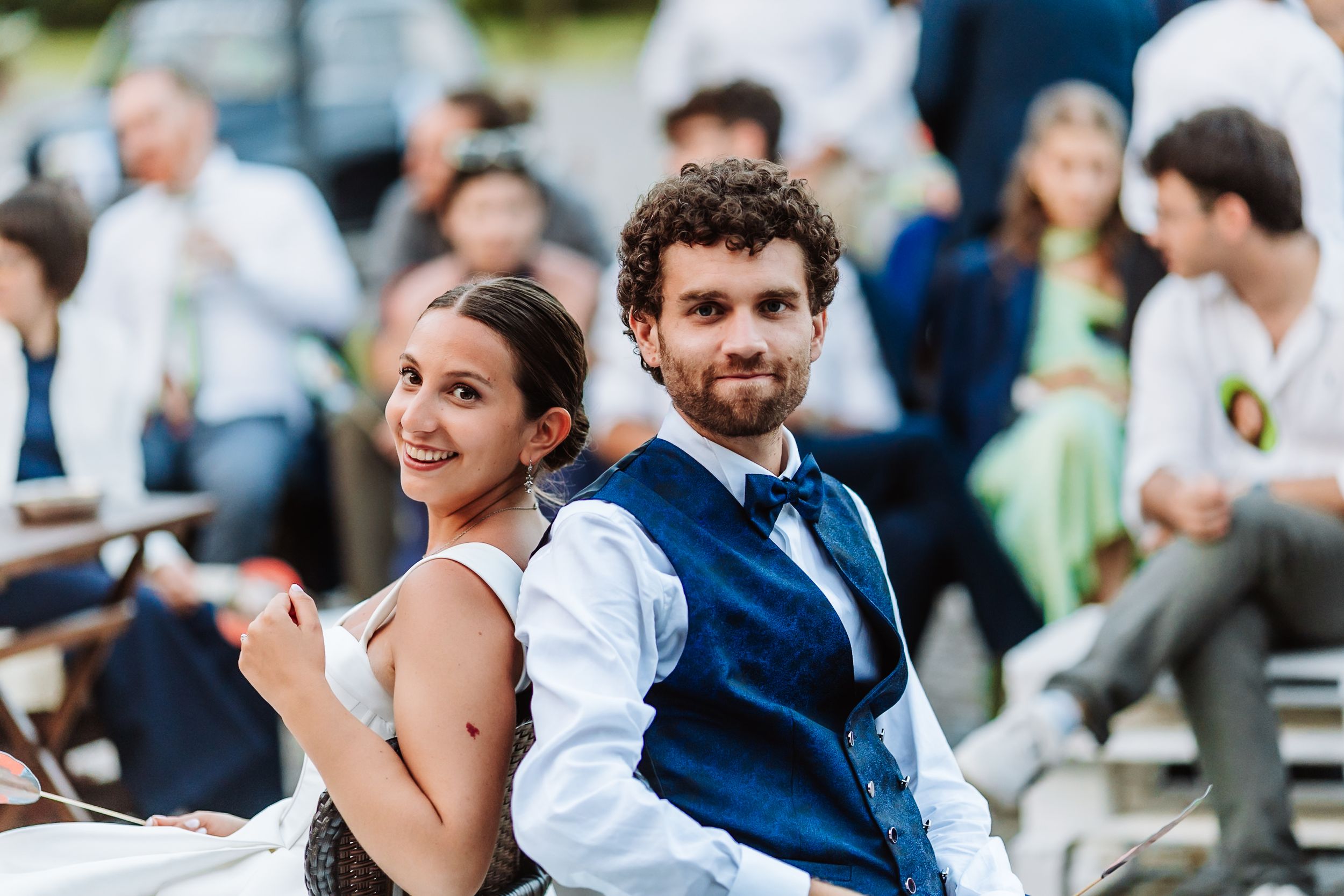 A smiling couple in formal attire poses back-to-back at an outdoor wedding reception.