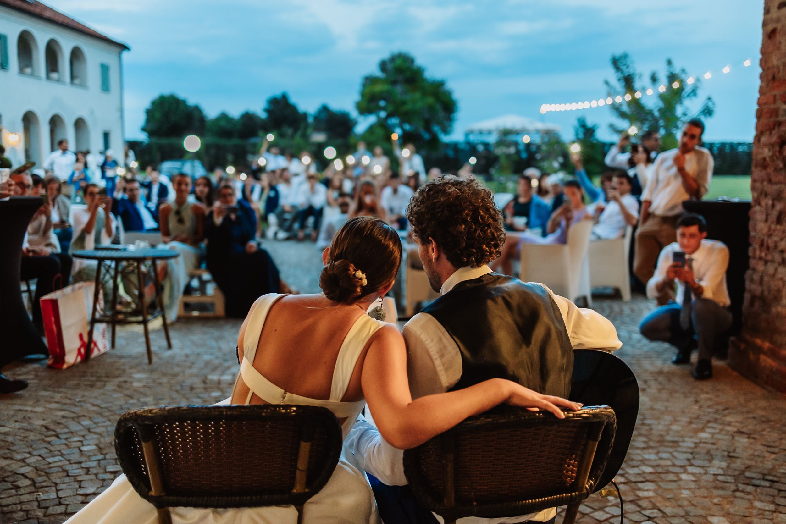 Bride and groom sit together at outdoor wedding reception while guests celebrate with sparklers at dusk.