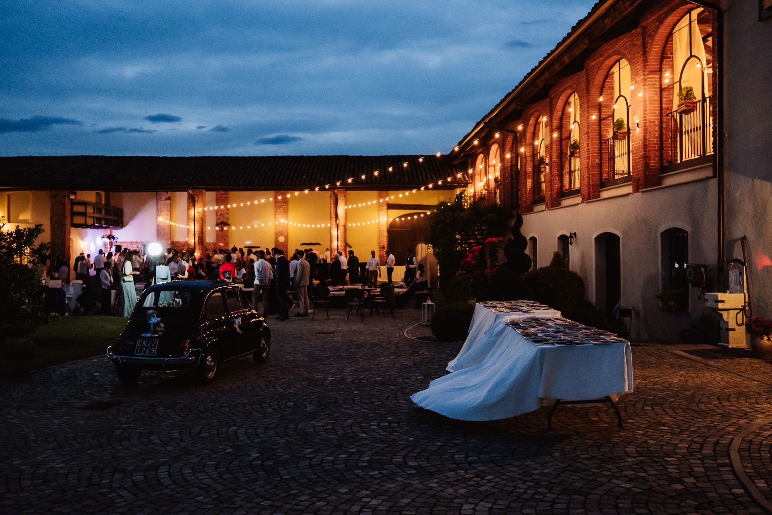Elegant outdoor evening event at a historic venue with string lights, cobblestone courtyard, and warmly lit arched windows.