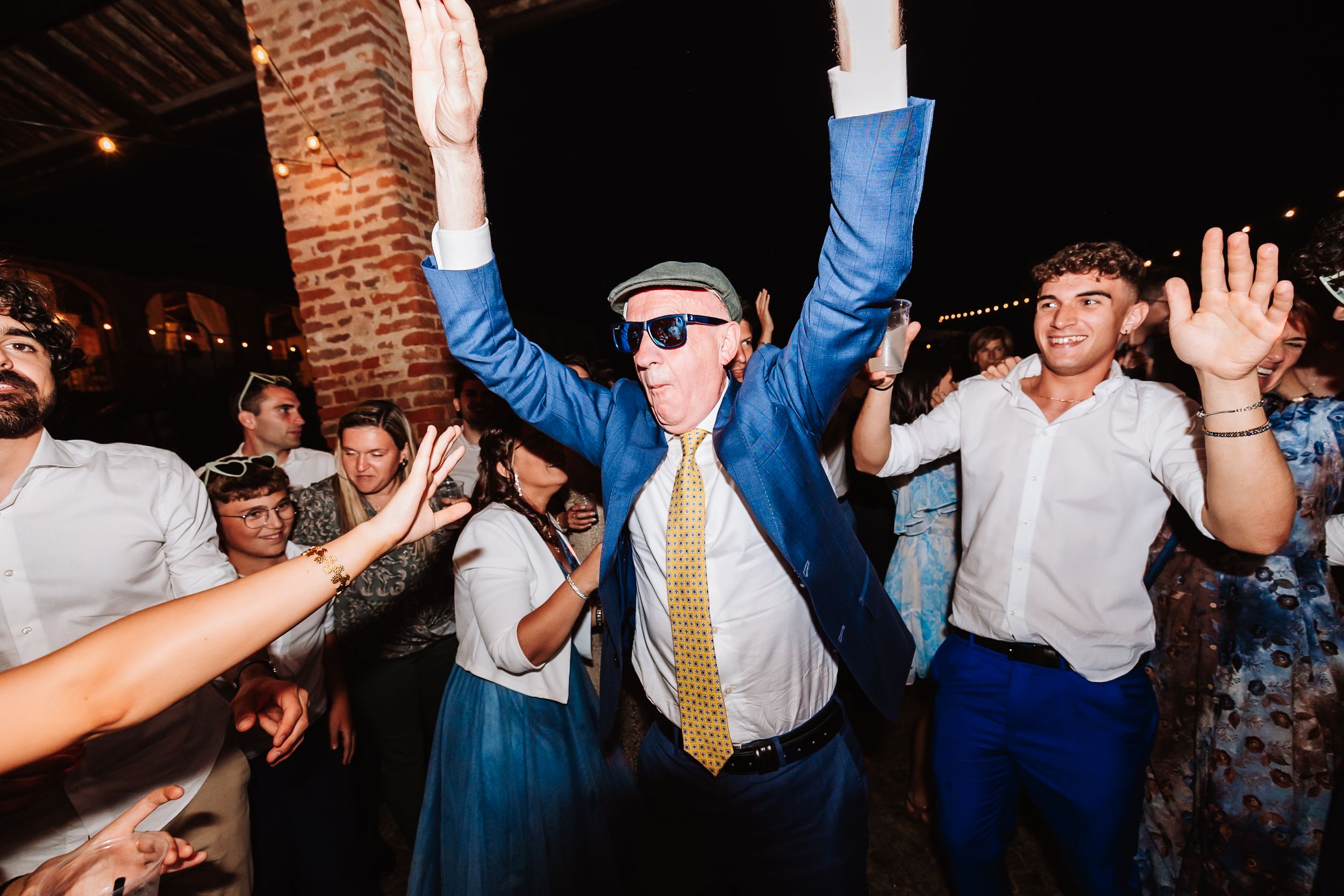 Elderly man in blue suit and flat cap raises arms joyfully on a lively wedding dance floor at night.