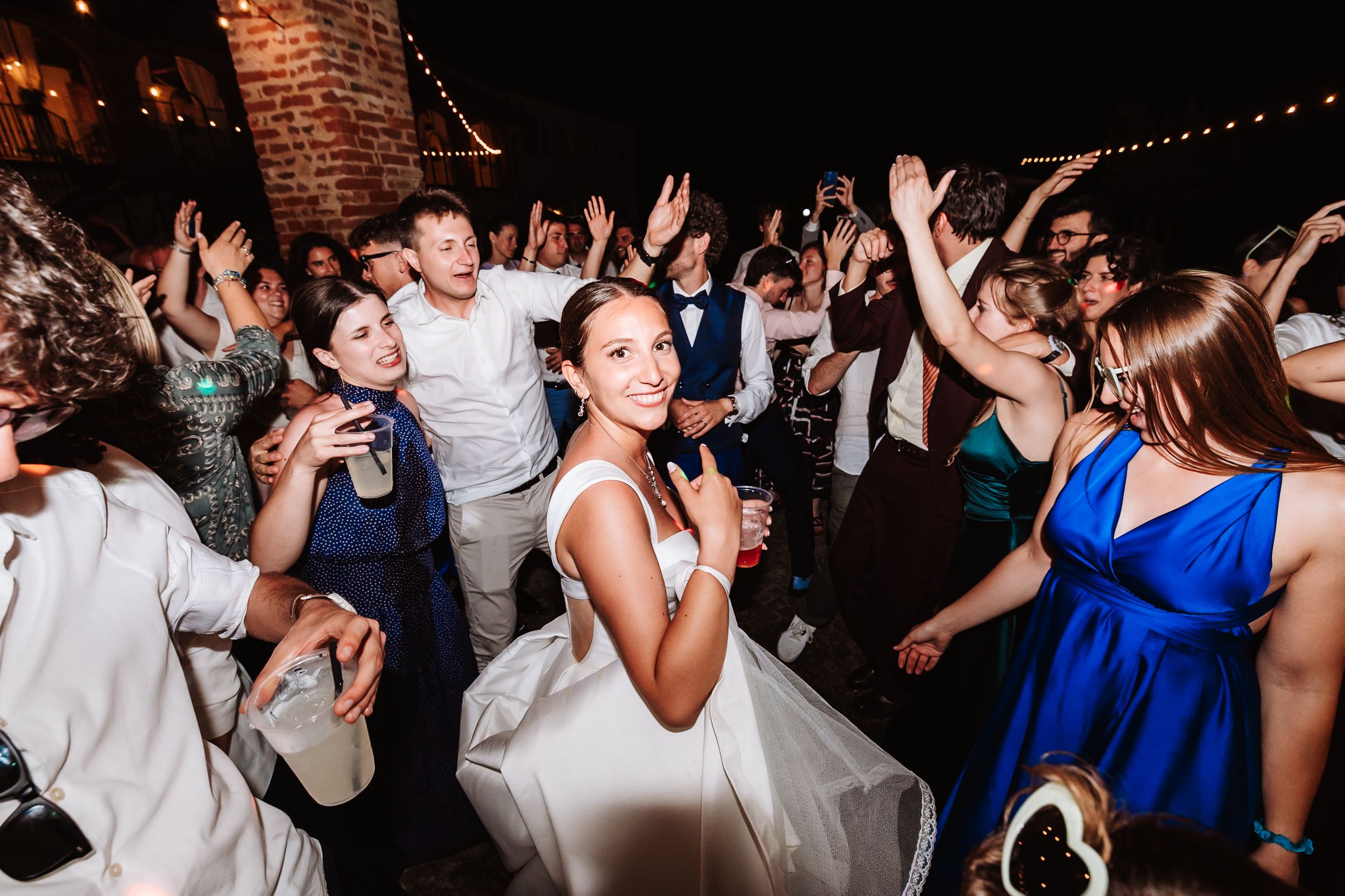 Bride in white wedding dress dancing joyfully with guests at a lively indoor wedding reception party.