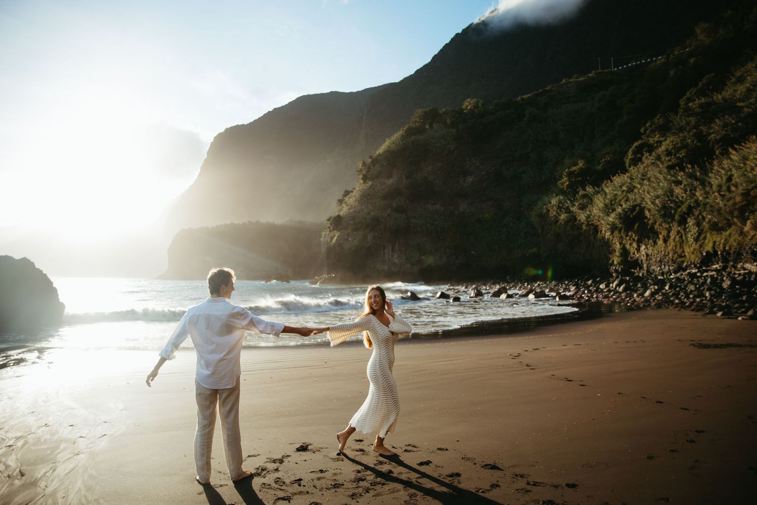 Couple dancing on a dramatic black sand beach at golden hour with towering green cliffs behind them.