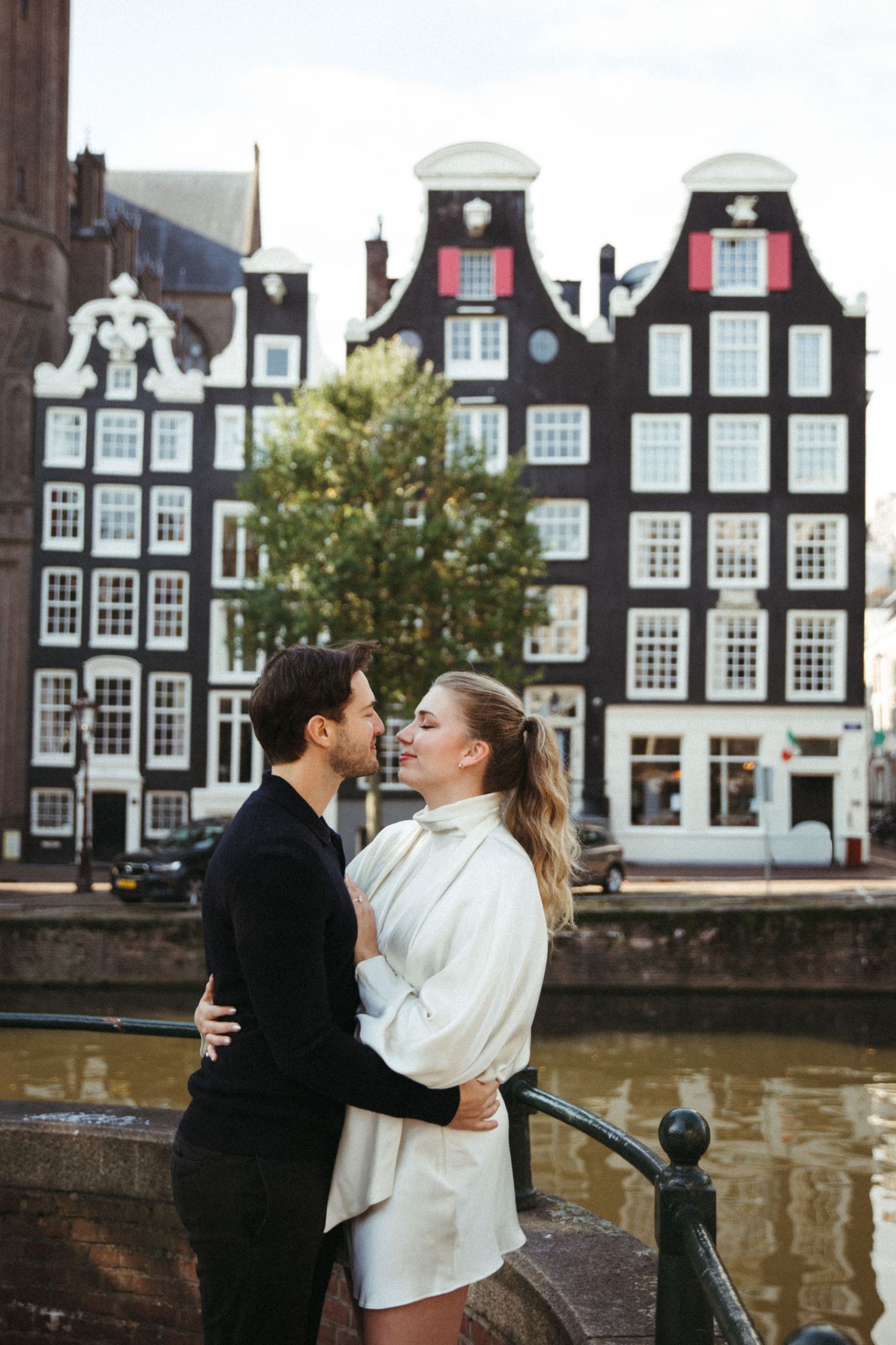 Romantic couple sharing a kiss on an Amsterdam canal bridge with iconic Dutch architecture behind them.