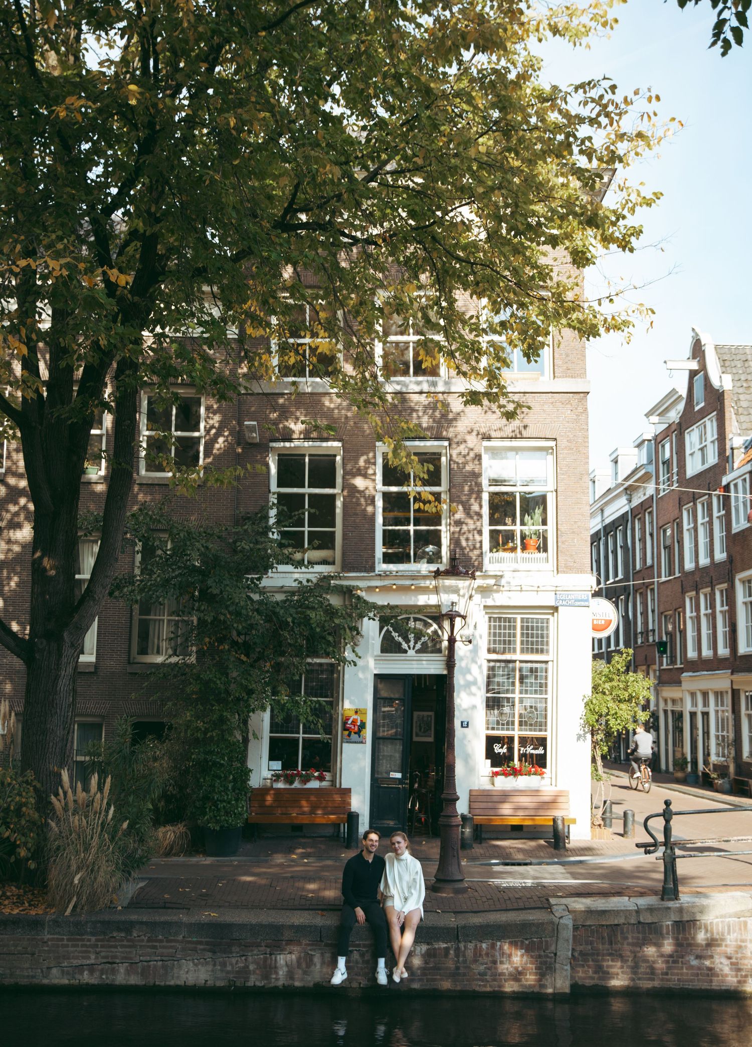 Couple sitting together outside a charming Amsterdam canal-side building surrounded by autumn trees.