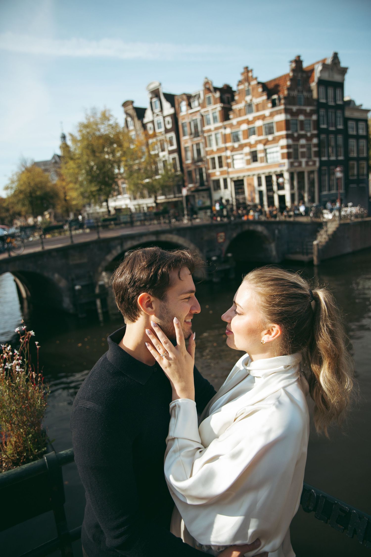 Loving couple embracing near an Amsterdam canal with historic bridge and traditional Dutch buildings.