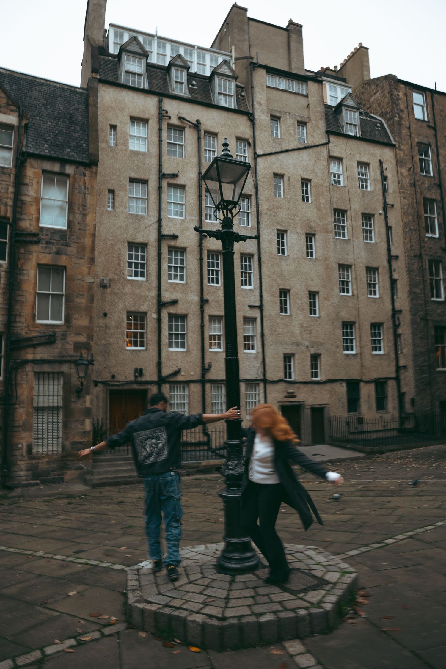 Couple playfully spinning around vintage lamppost in historic Edinburgh courtyard.