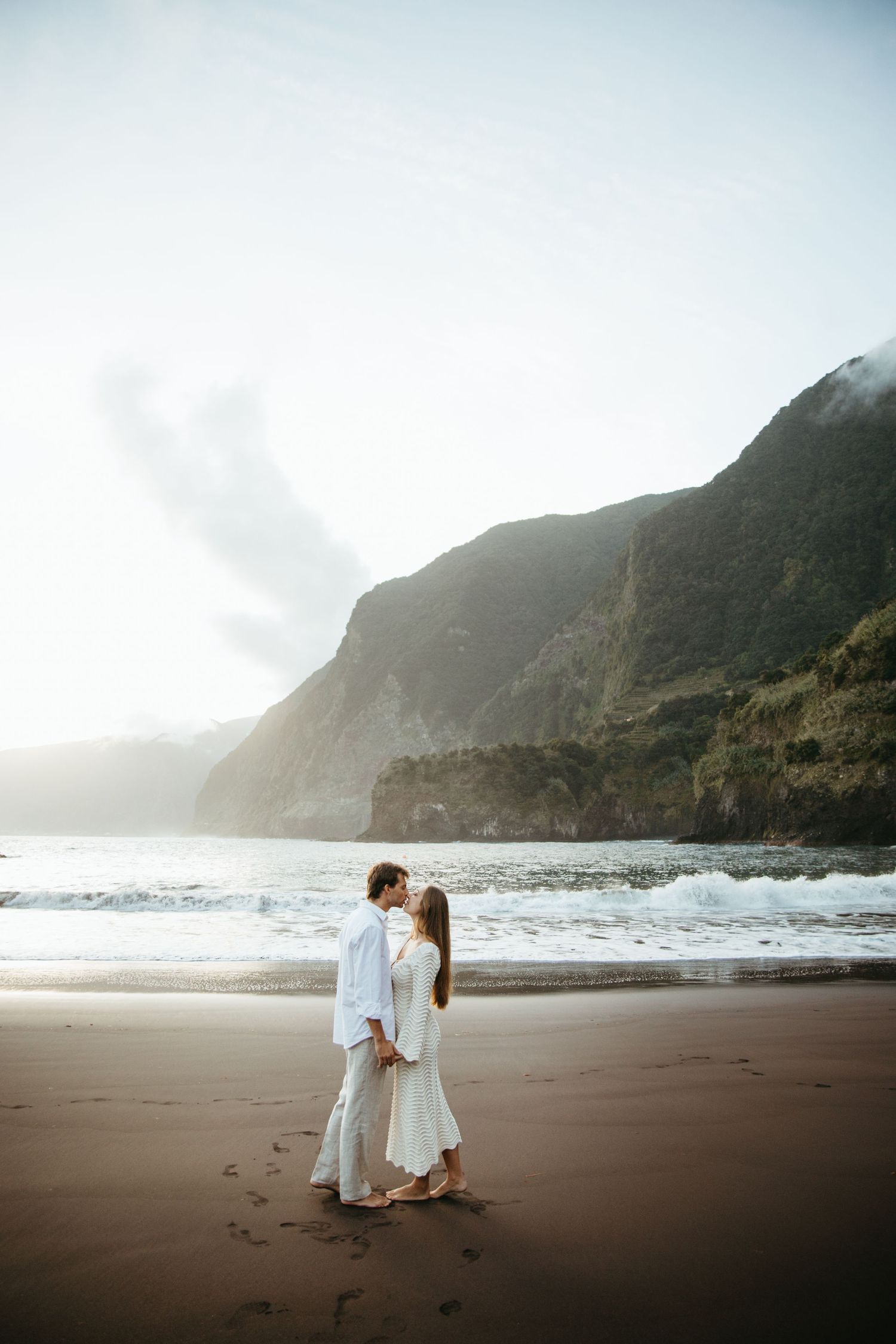 Couple in white attire sharing a kiss on dark sand beach with dramatic cliffs.