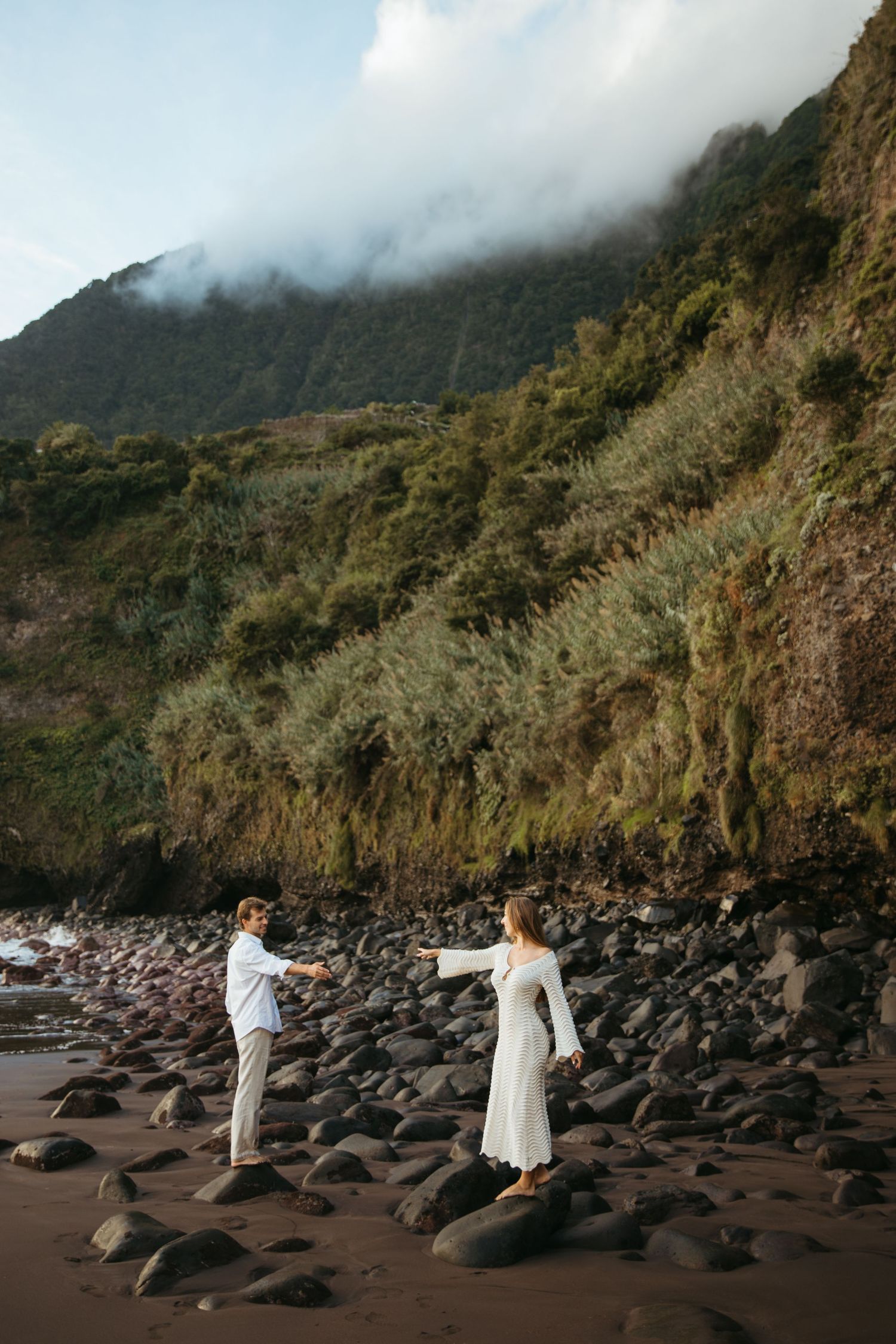 Couple in white holding hands on rocky volcanic beach with misty green cliffs.