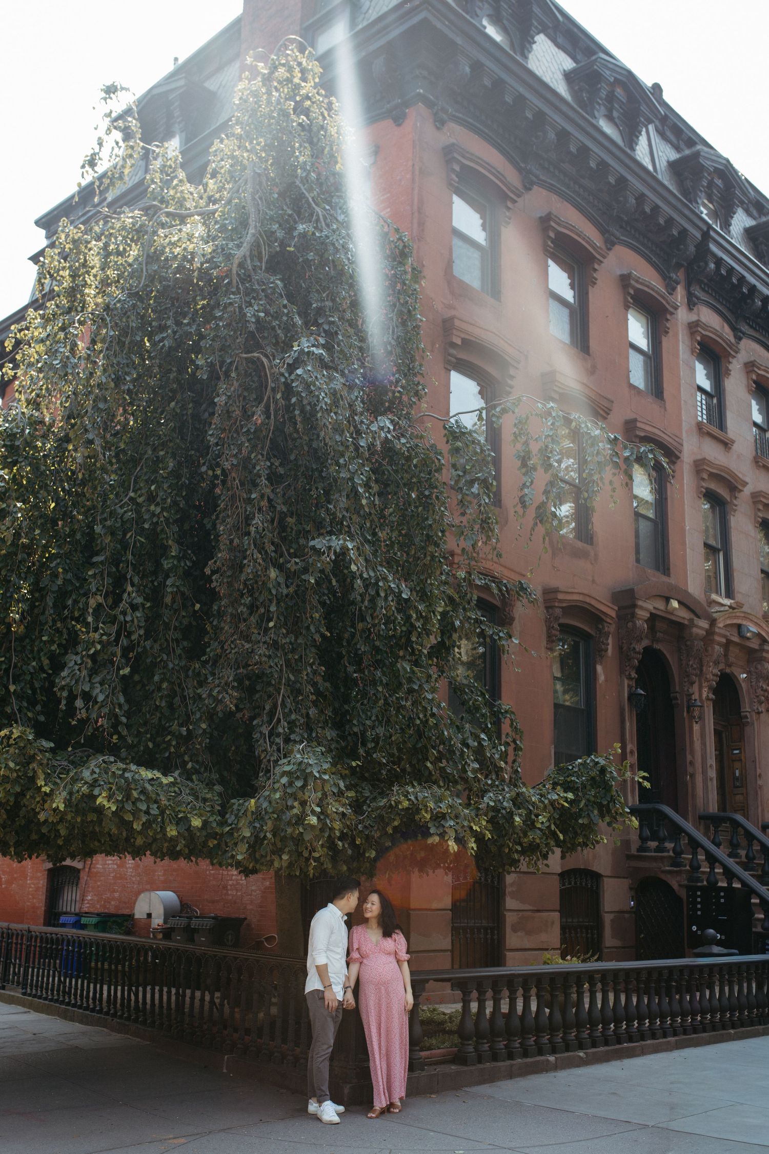 Couple holding hands in front of a ivy-covered brownstone building on a sunny New York City street.