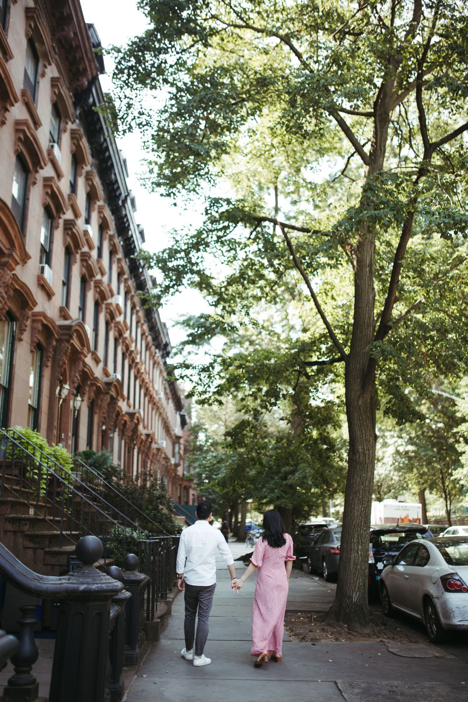 Couple walking hand in hand down a sunlit Brooklyn street lined with classic brownstone buildings.