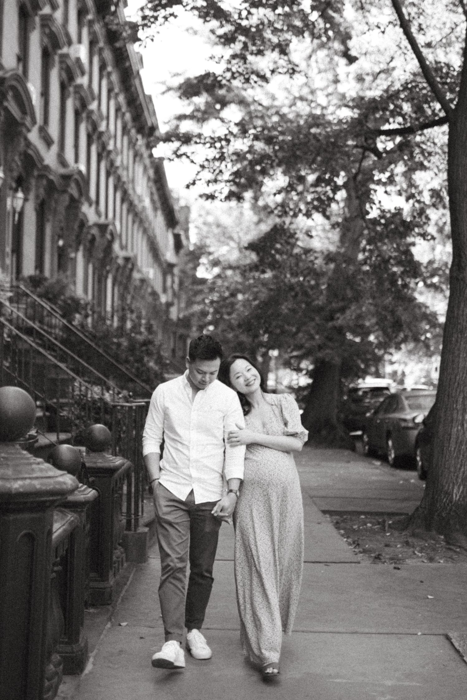 Black and white photo of a couple walking arm in arm along a tree-lined Brooklyn brownstone sidewalk.