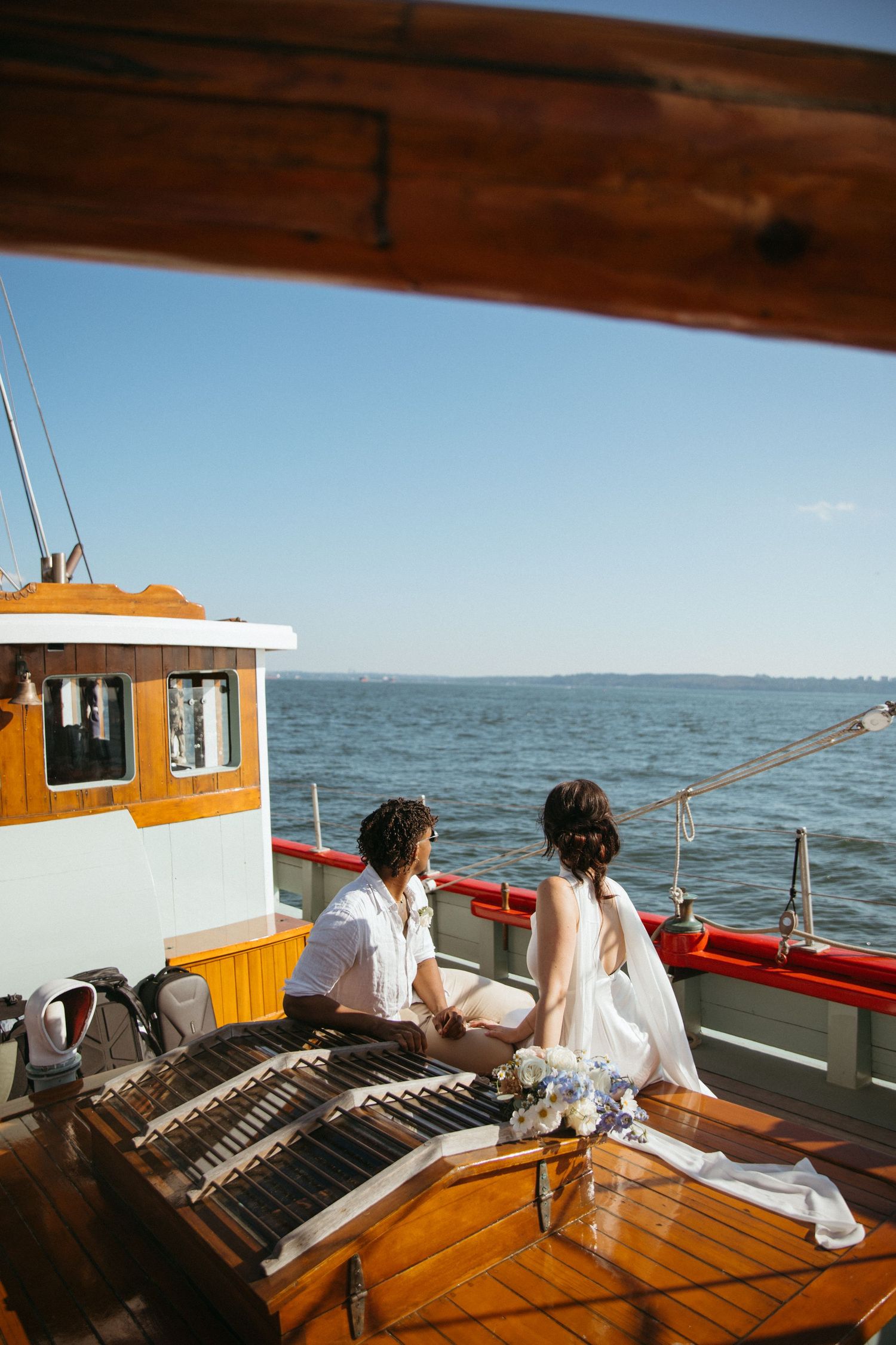 Bride and groom sitting on classic wooden boat deck overlooking open blue water.