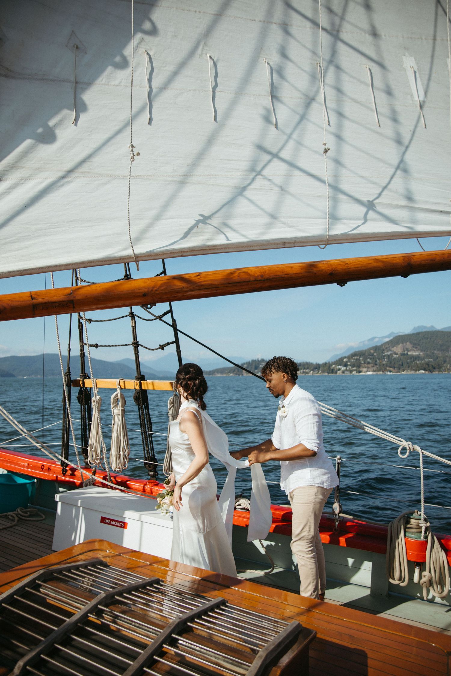 Bride and groom standing on sailboat deck with white sails and mountain backdrop.